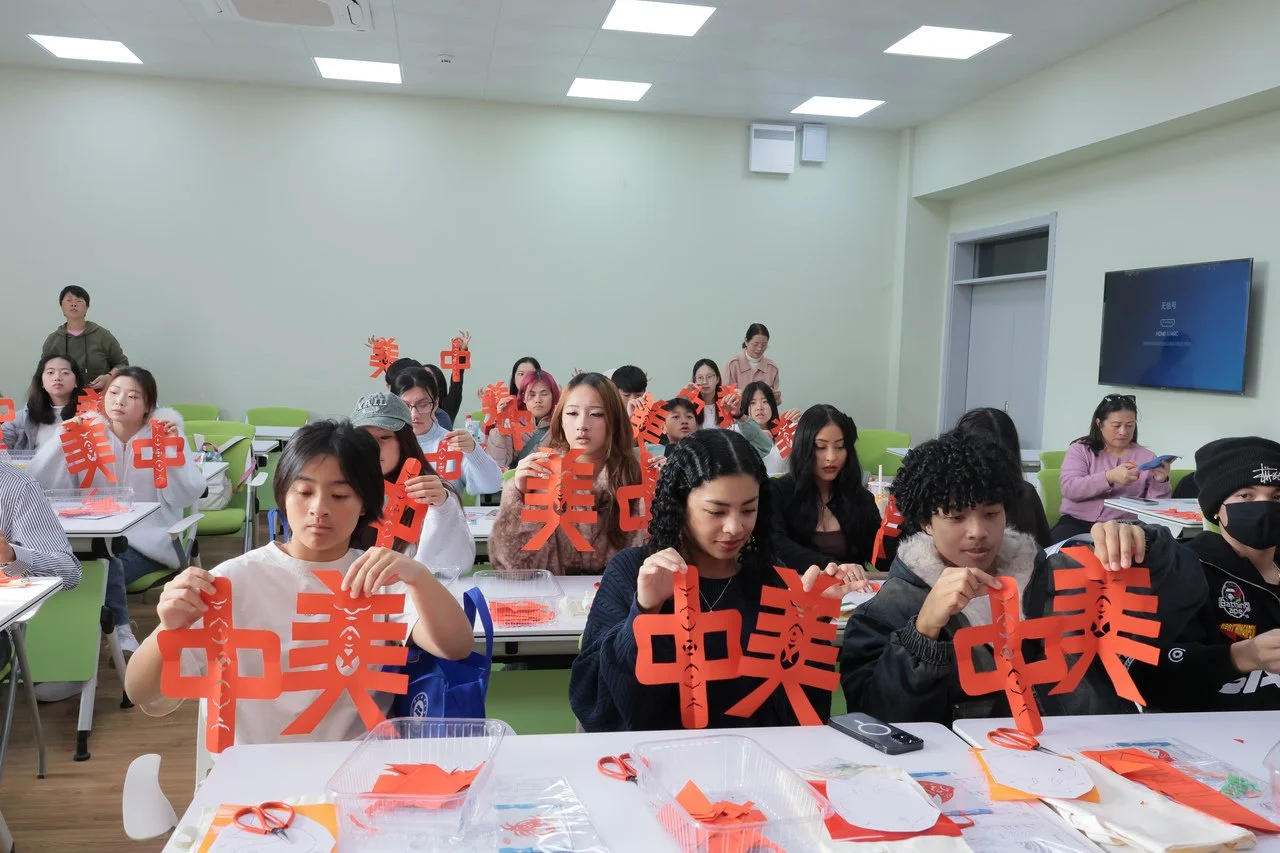 Group of people sitting at tables in a classroom, holding up red paper cutouts with Chinese characters, participating in a craft activity.