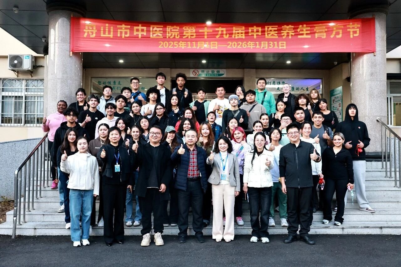 Group of diverse people standing on steps in front of a building, posing for a photo during a cultural event at a Chinese medical hospital, with a red banner overhead.