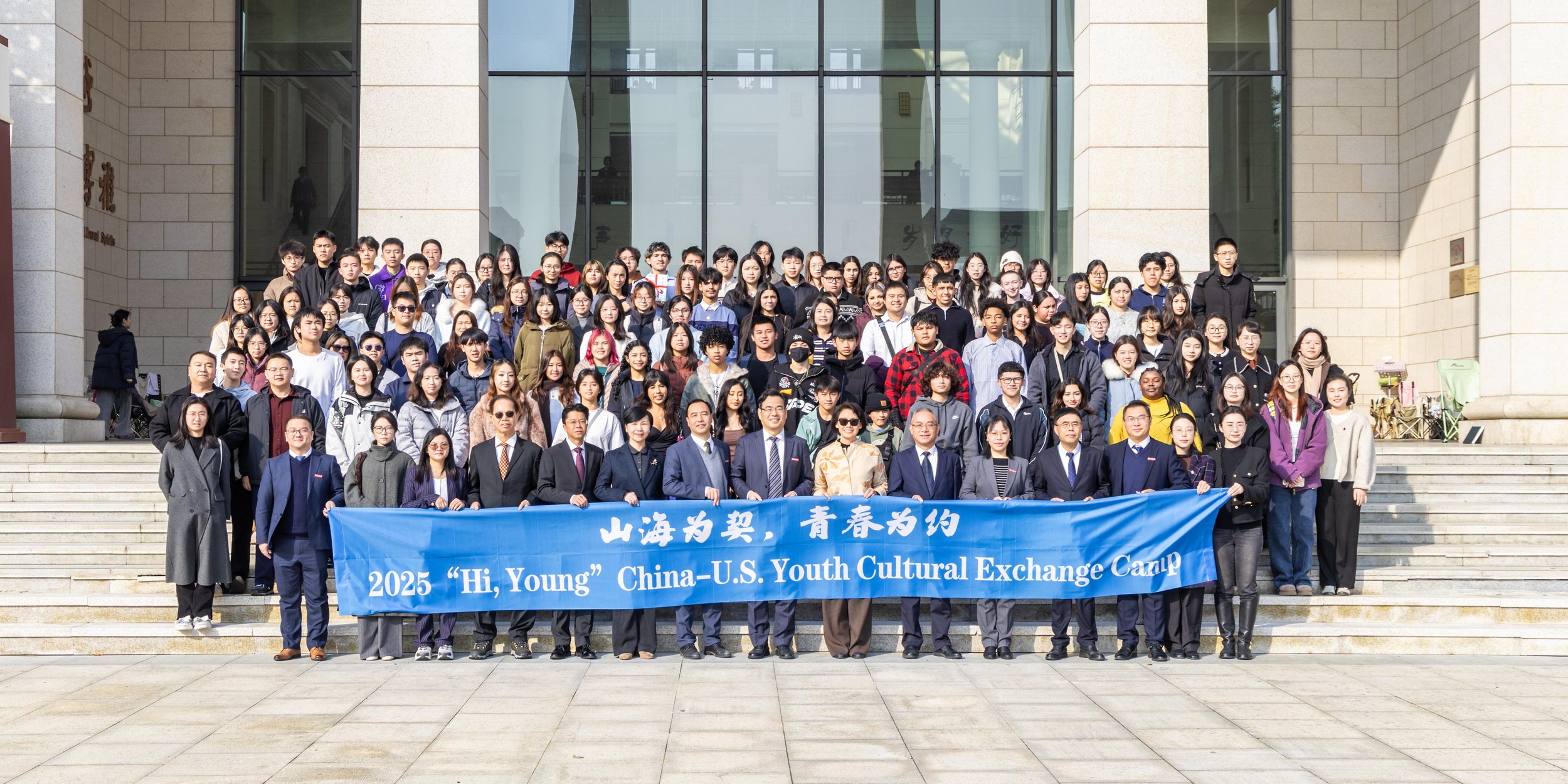 Group photo of diverse young people and adults standing on steps outside a building with glass front, holding a blue banner that reads '2025 Hi, Young China-U.S. Youth Cultural Exchange Camp'.