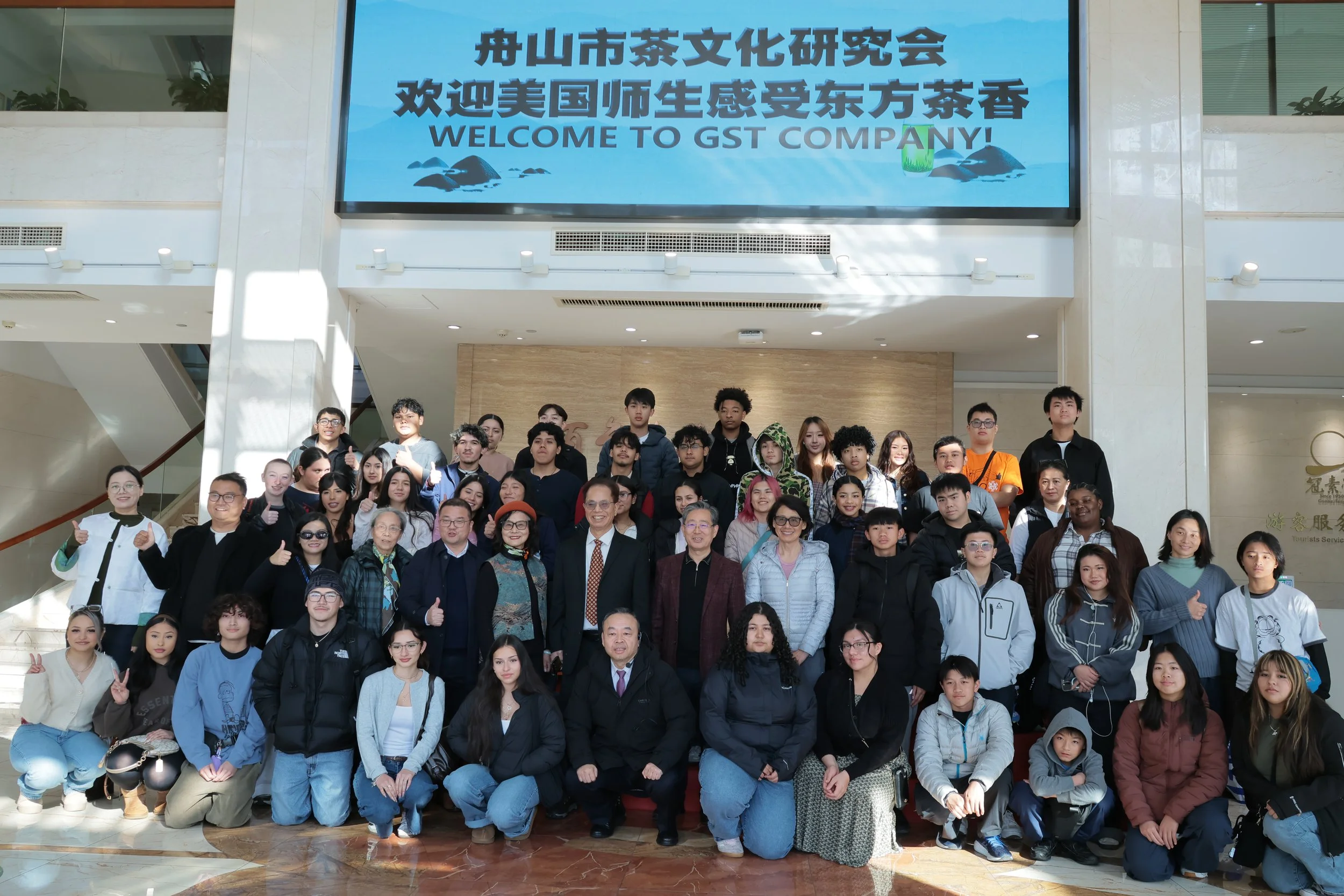Group of people posing for a photo in a spacious indoor area with a large digital sign overhead that says 'Welcome to GST Company' in English and Chinese, with a scenic background of mountains, rocks, and water.