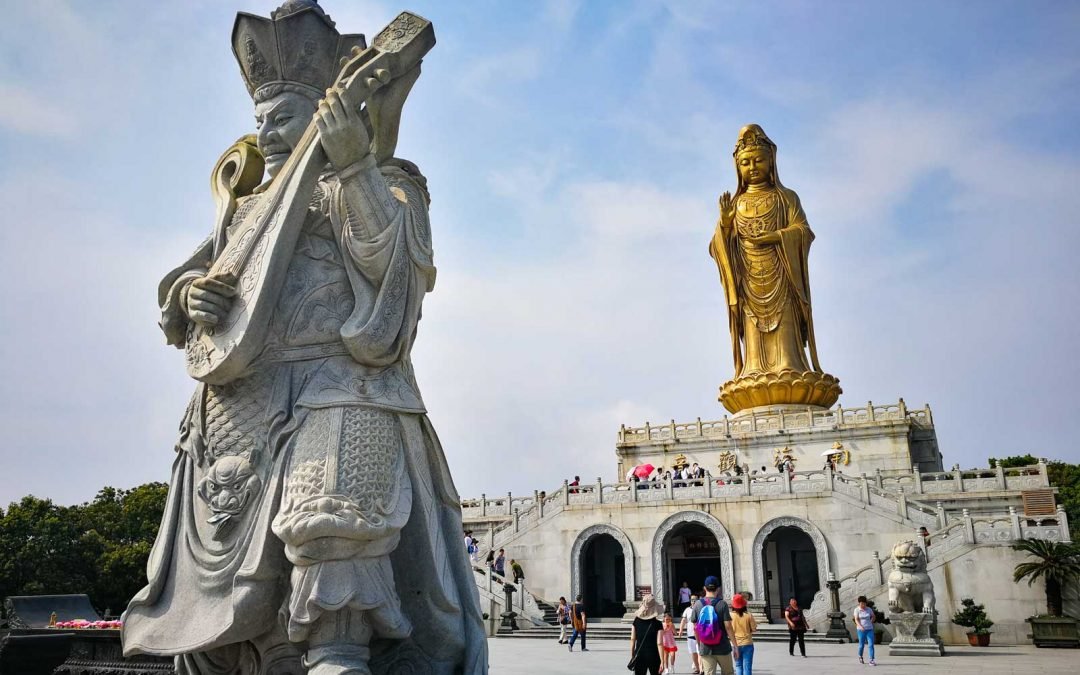 A large marble statue of a traditional Chinese warrior playing a stringed instrument in the foreground, with a tall golden statue of a Buddhist figure on a platform in the background, and visitors walking around.