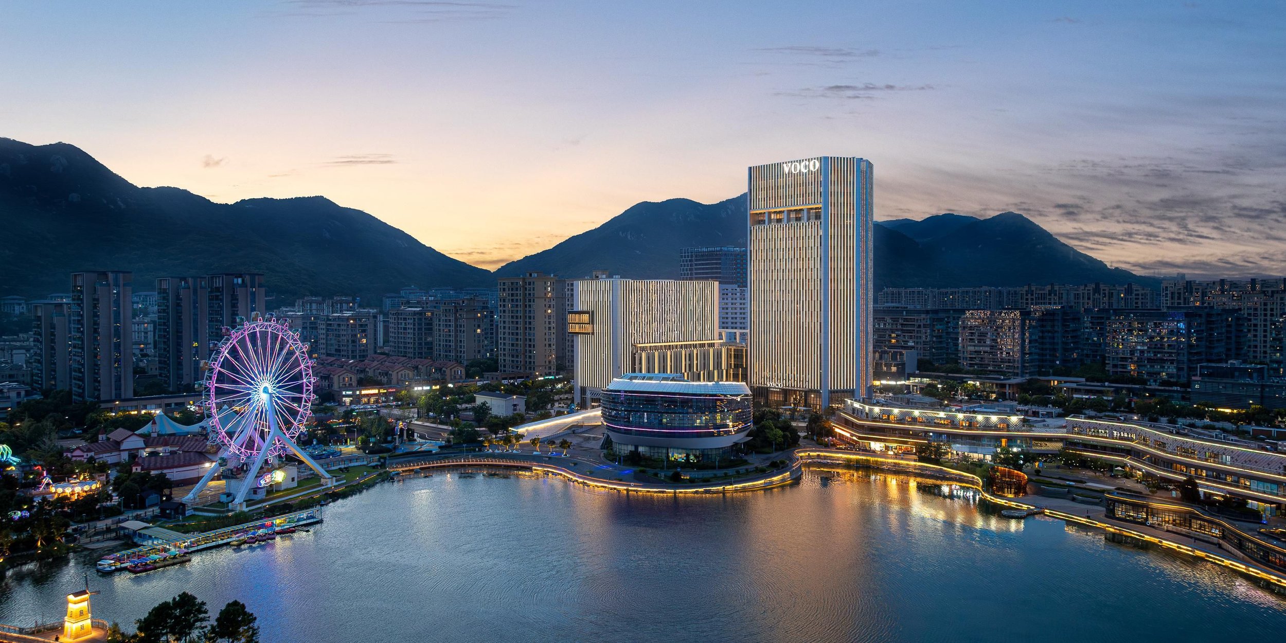 City skyline at sunset with tall modern buildings, a large Ferris wheel illuminated in pink, and a body of water reflecting city lights, mountain range in the background.