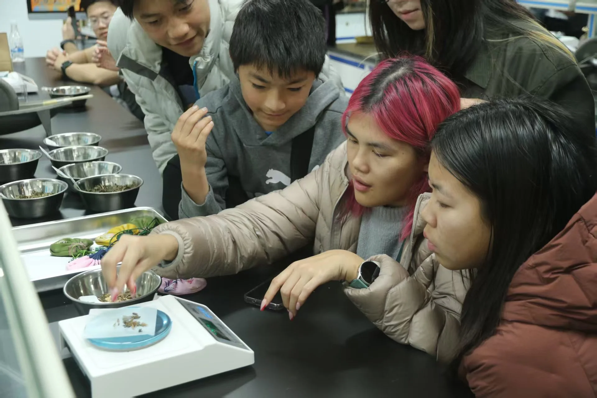A group of people gathered around a table, closely observing a woman with pink hair who is using a scale to weigh small objects, likely for a science experiment.