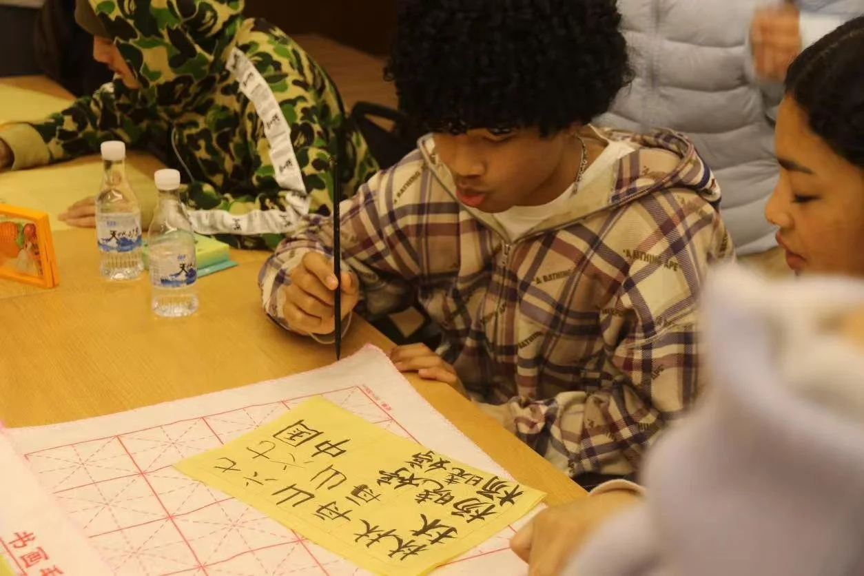 Three young people sitting at a table, focusing on writing or reading. The table has water bottles, notebooks, and a sheet with Japanese or Chinese characters. One person is in the foreground with curly hair and plaid hoodie, another in the backgroun
