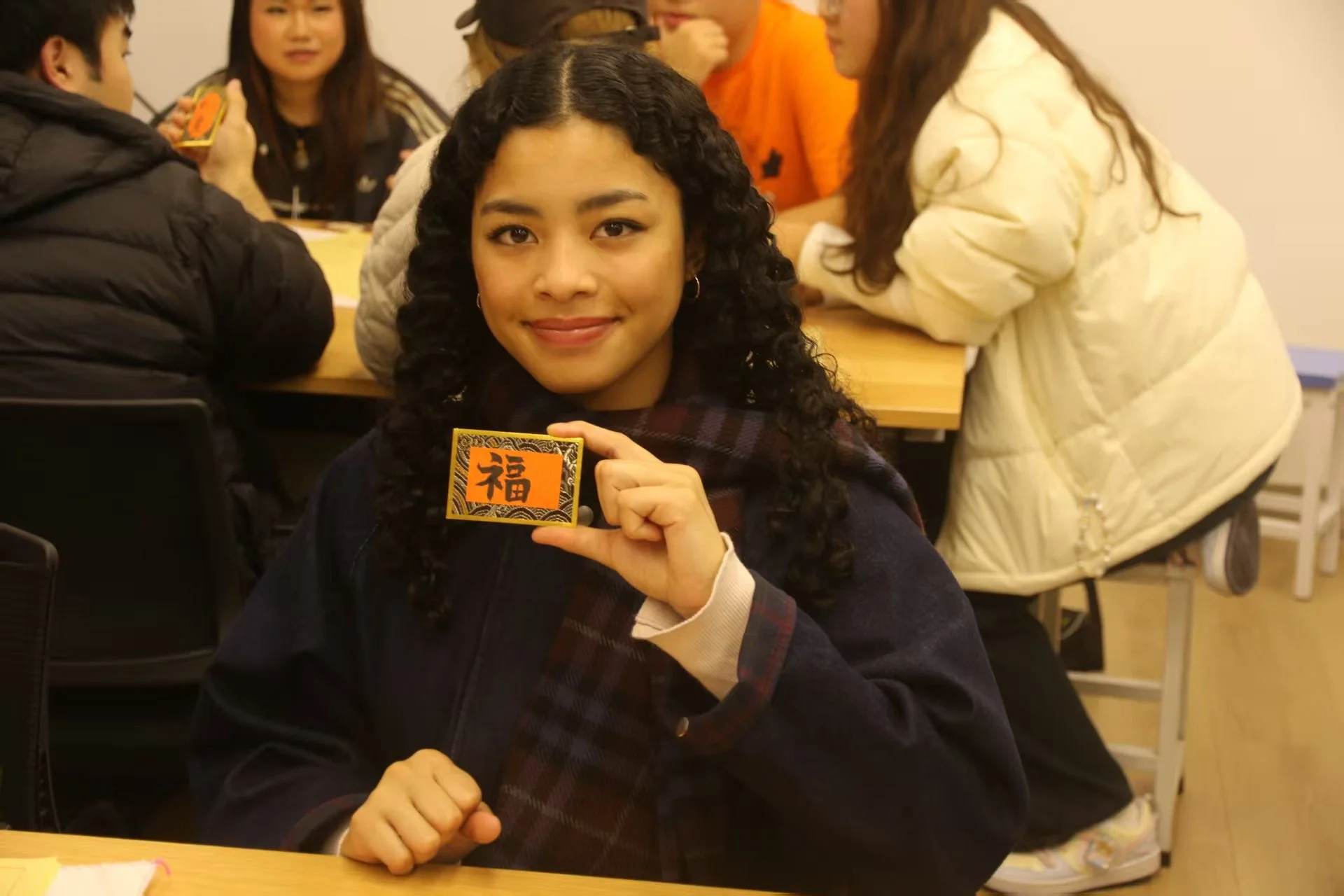 A young woman with curly hair smiling and holding a red envelope with a Chinese character, sitting at a table with other people in the background.