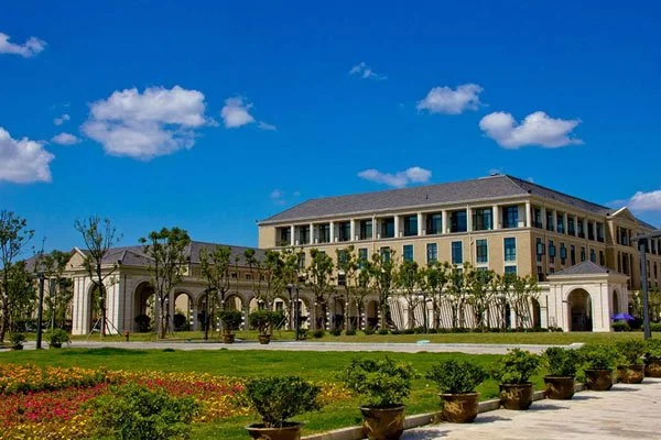 Large modern building with multiple windows, surrounded by a landscaped garden with potted plants, under a bright blue sky with some clouds.