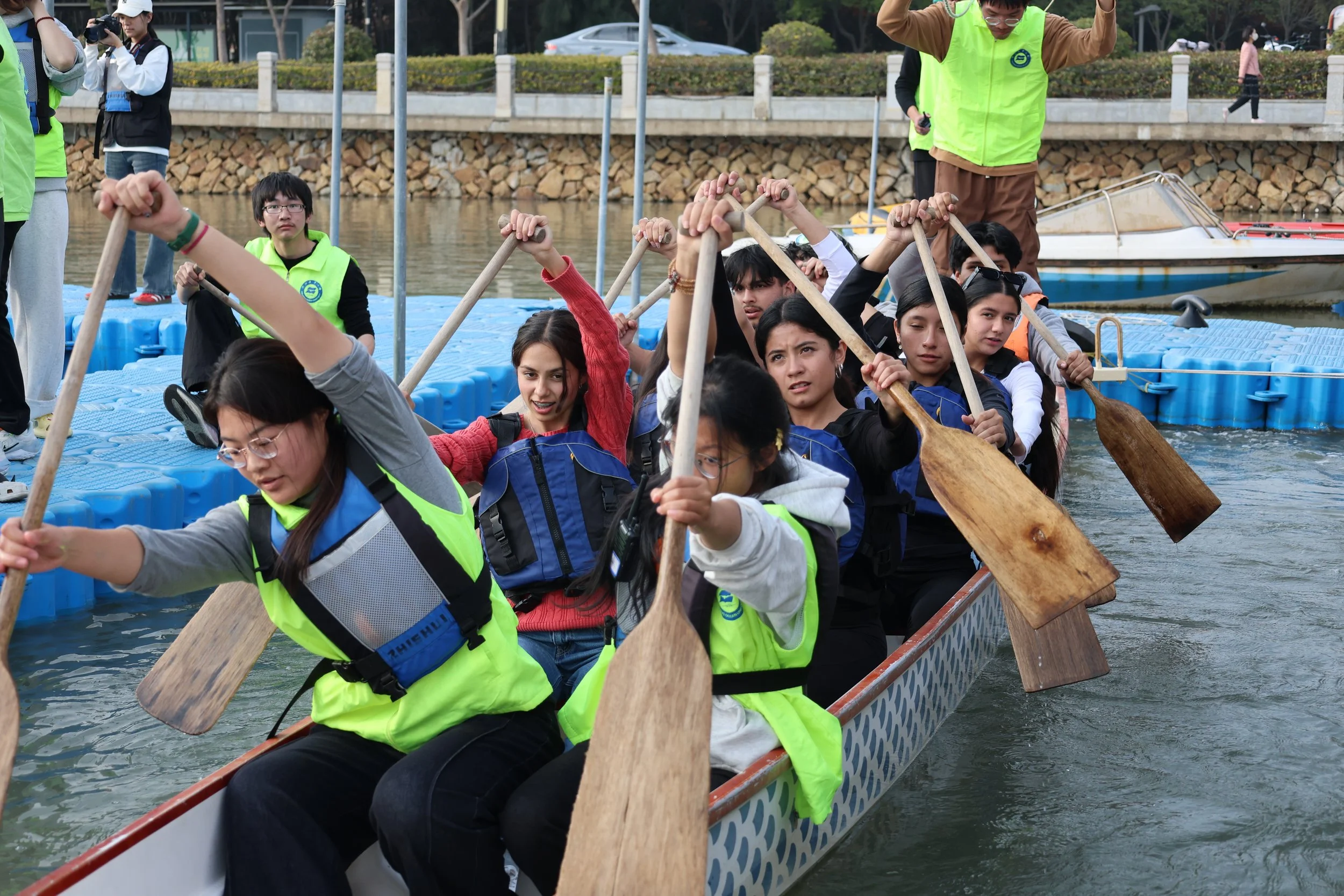 A group of people participating in a dragon boat race, paddling vigorously on a boat with advance planning and coordination, on a river with a rocky shore and other boats in the background.