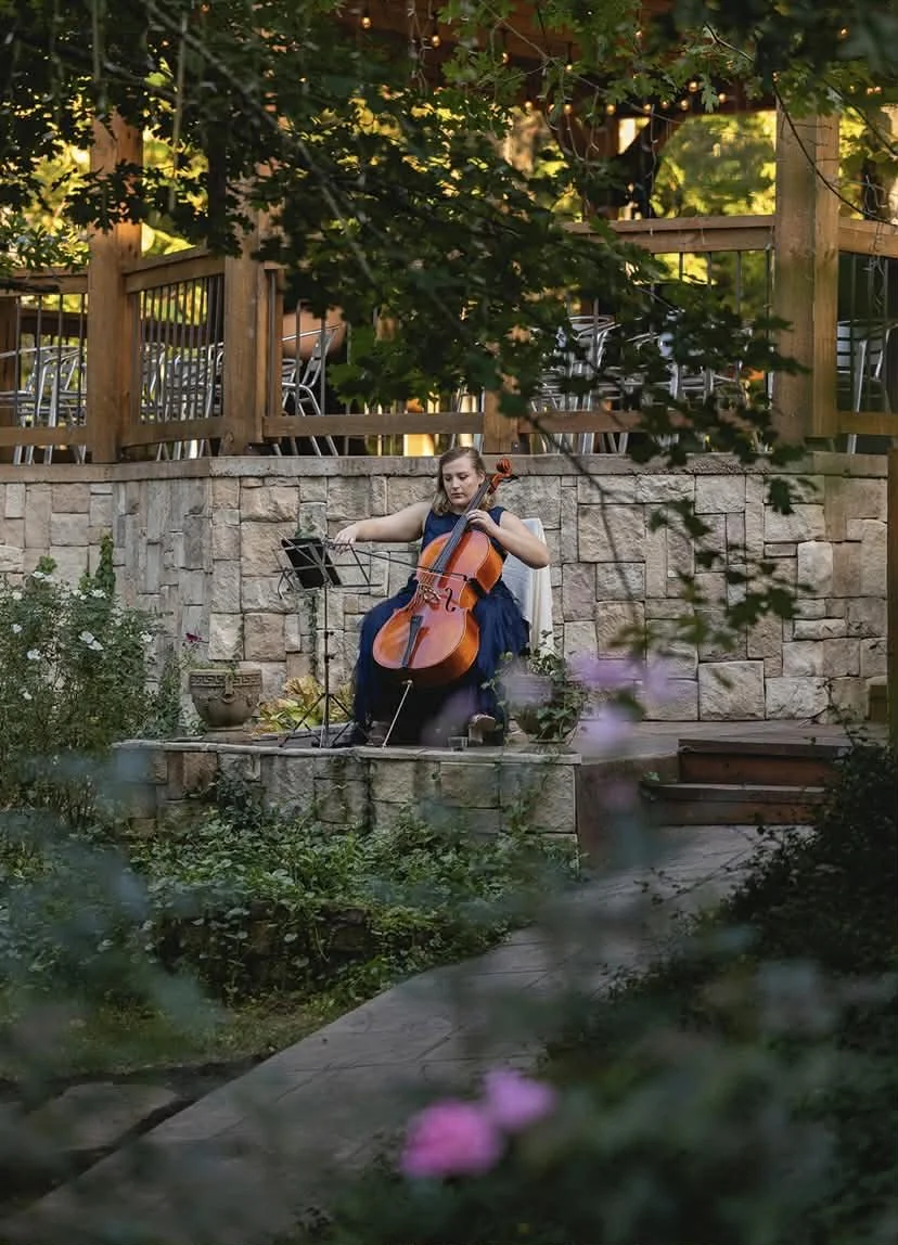 Carlyn playing for an outdoor wedding in a garden area surrounded by greenery and flowers, with a stone wall and a wooden deck above.