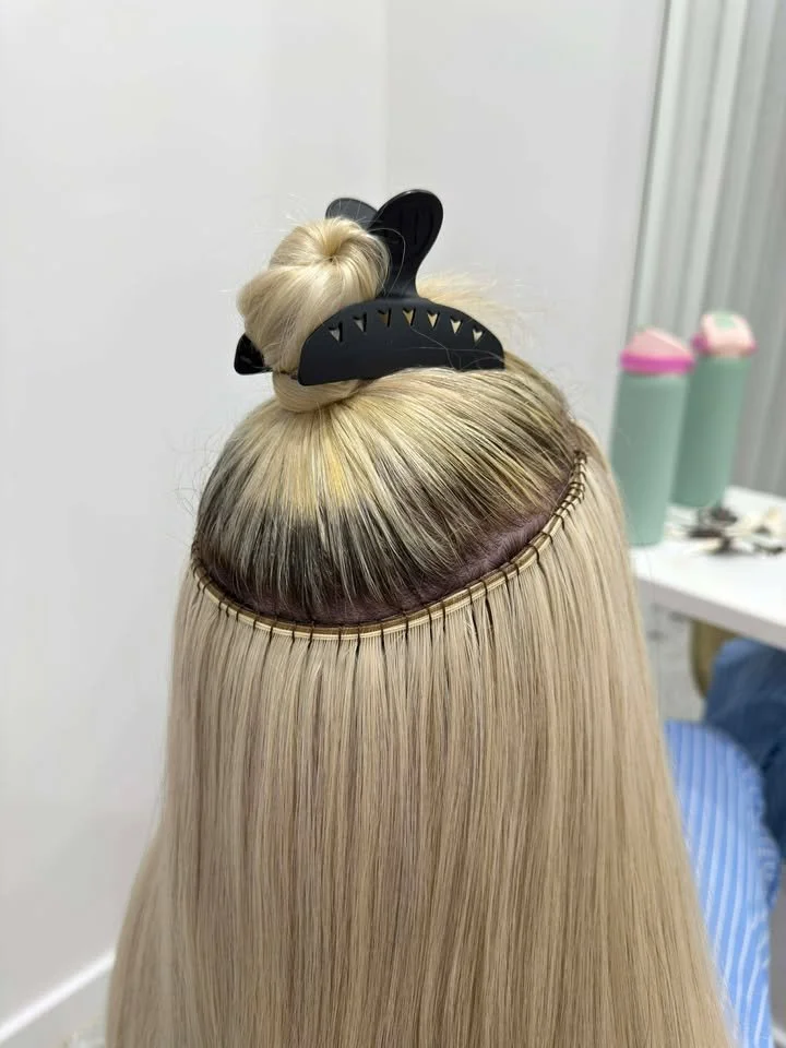 Blonde hair with a section at the crown lifted and held with a black hair clip, showing a lace front wig or hair extension installation process.