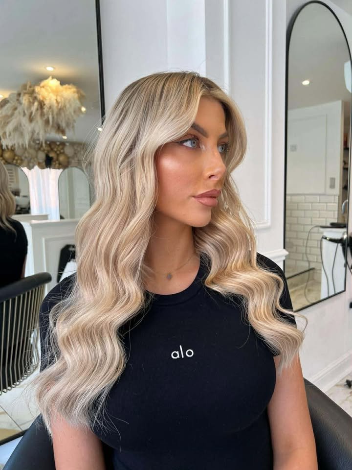A woman with long, wavy blonde hair styled in loose waves, wearing a black shirt with the word 'alo' on it, sitting in a salon with mirrors and salon equipment visible in the background.