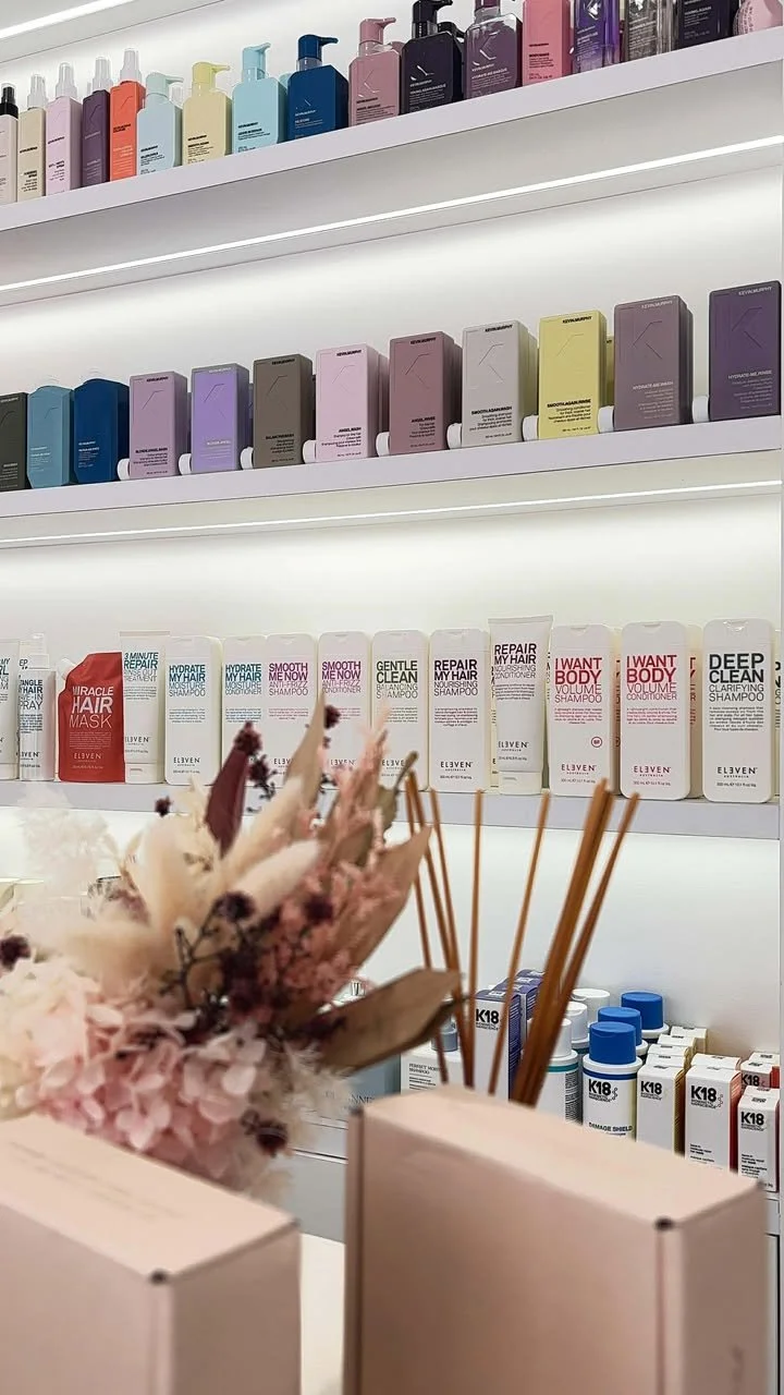 Display of various hair care and styling products on shelves, including bottles and boxes in different colors, with dried flowers in the foreground.