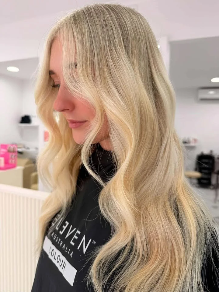 A woman with long, wavy blonde hair and bangs, looking downward in a salon.