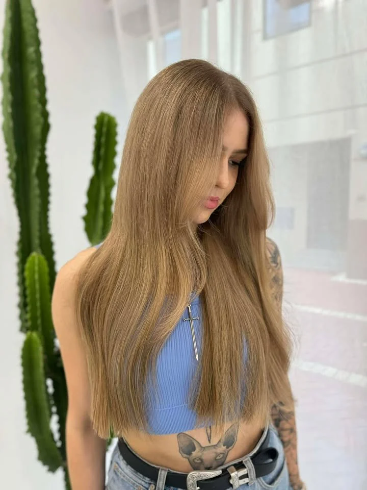 A young woman with long, straight, blonde hair, wearing a blue sleeveless top and tattoos on her arms and stomach, standing indoors near green cacti.
