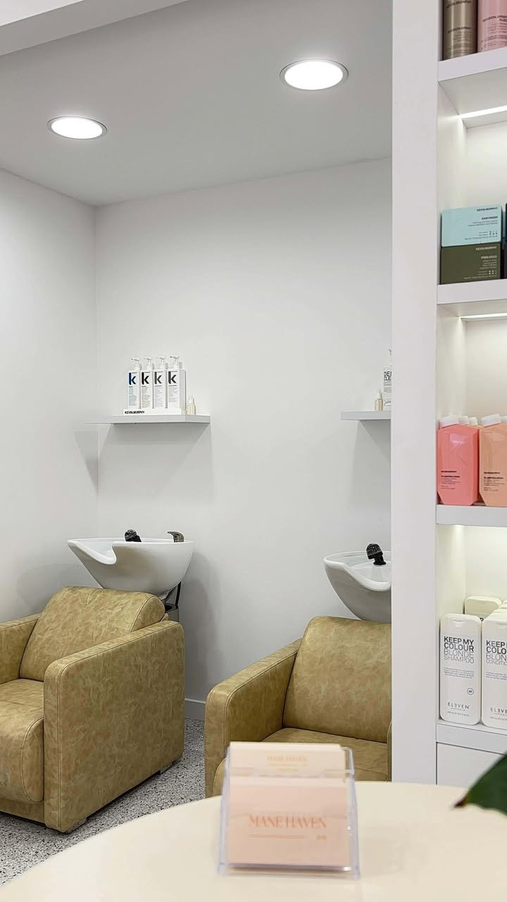 Salon with two beige chairs, two white shampoo sinks, shelves with hair products, and a table with a stack of pink and white hair color swatches.