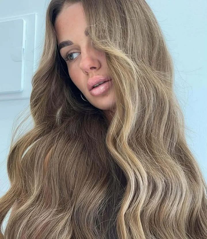 Close-up of a woman with long, wavy, light brown hair and makeup, looking slightly to the side.