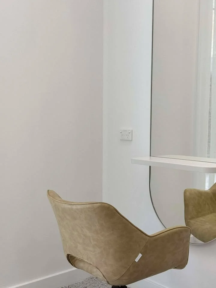 A modern interior space with a tan suede lounge chair in the foreground, and a white wall with an electrical outlet and a large mirror reflected on the right.