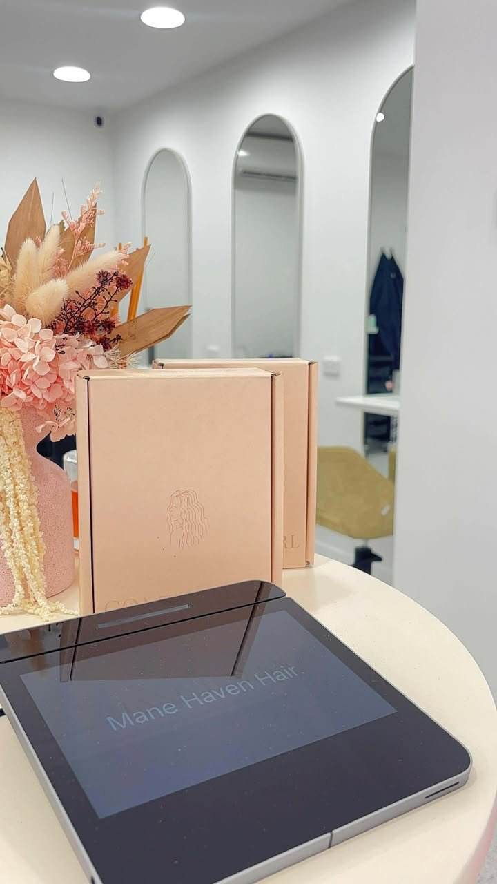 A digital device on a white table displays the text "Mane Haven Hair." There are pink and beige boxes and flowers nearby, with a white wall and arched mirrors in the background.