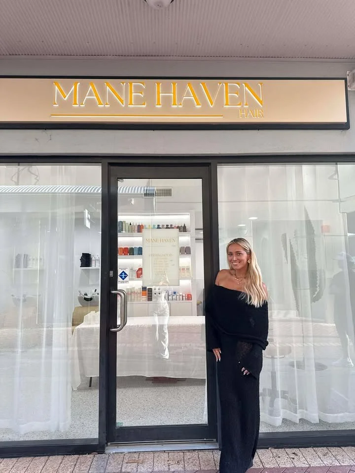 Woman standing outside Mane Haven Hair salon, smiling, wearing black off-the-shoulder sweater and black pants.