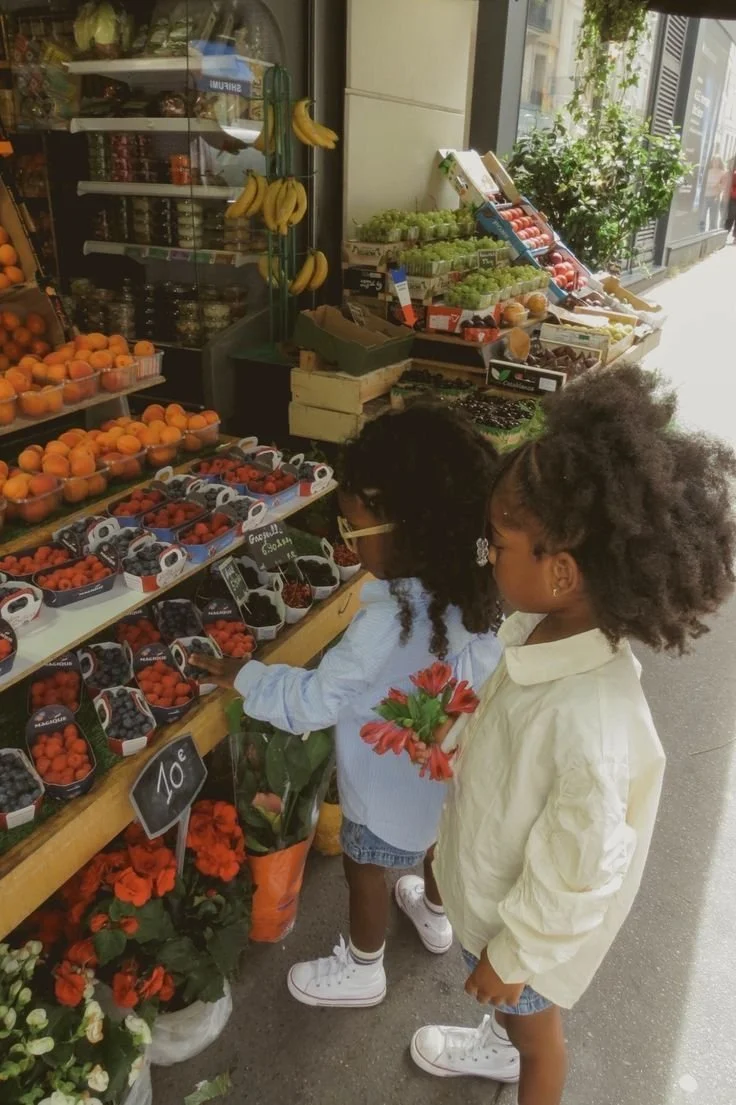 Two young girls shopping for berries at an outdoor fruit stand, with bananas, grapes, peaches, and other fruits displayed.