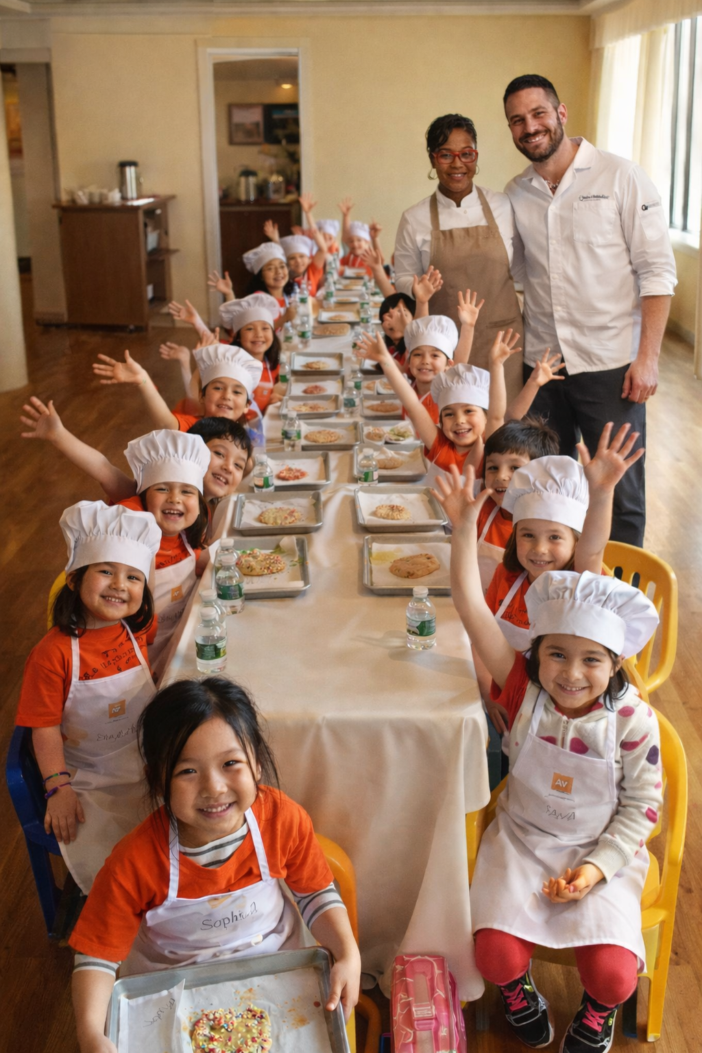 Children in chef hats and aprons sitting at a long table with cookie ingredients, smiling and raising their hands, with two adult chefs standing at the head of the table.