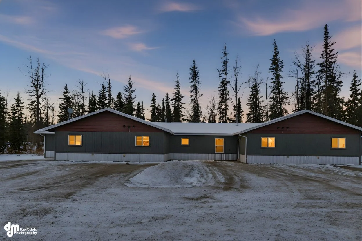 A single-story house with dark gray and red exterior, illuminated windows, surrounded by a snow-covered yard and trees in the background during dusk.