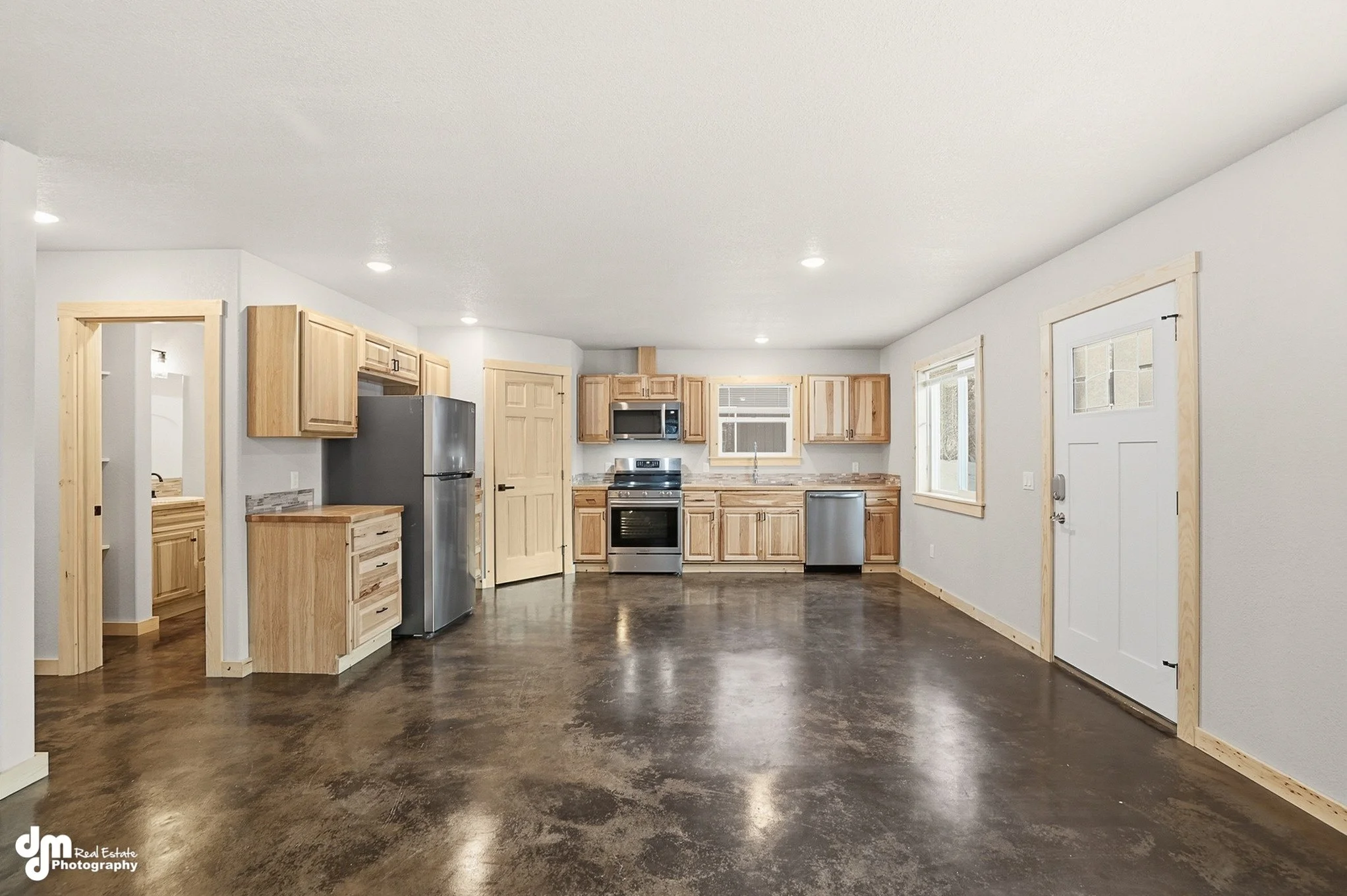 Empty kitchen with natural wood cabinets, stainless steel appliances, white walls, and a dark polished concrete floor.