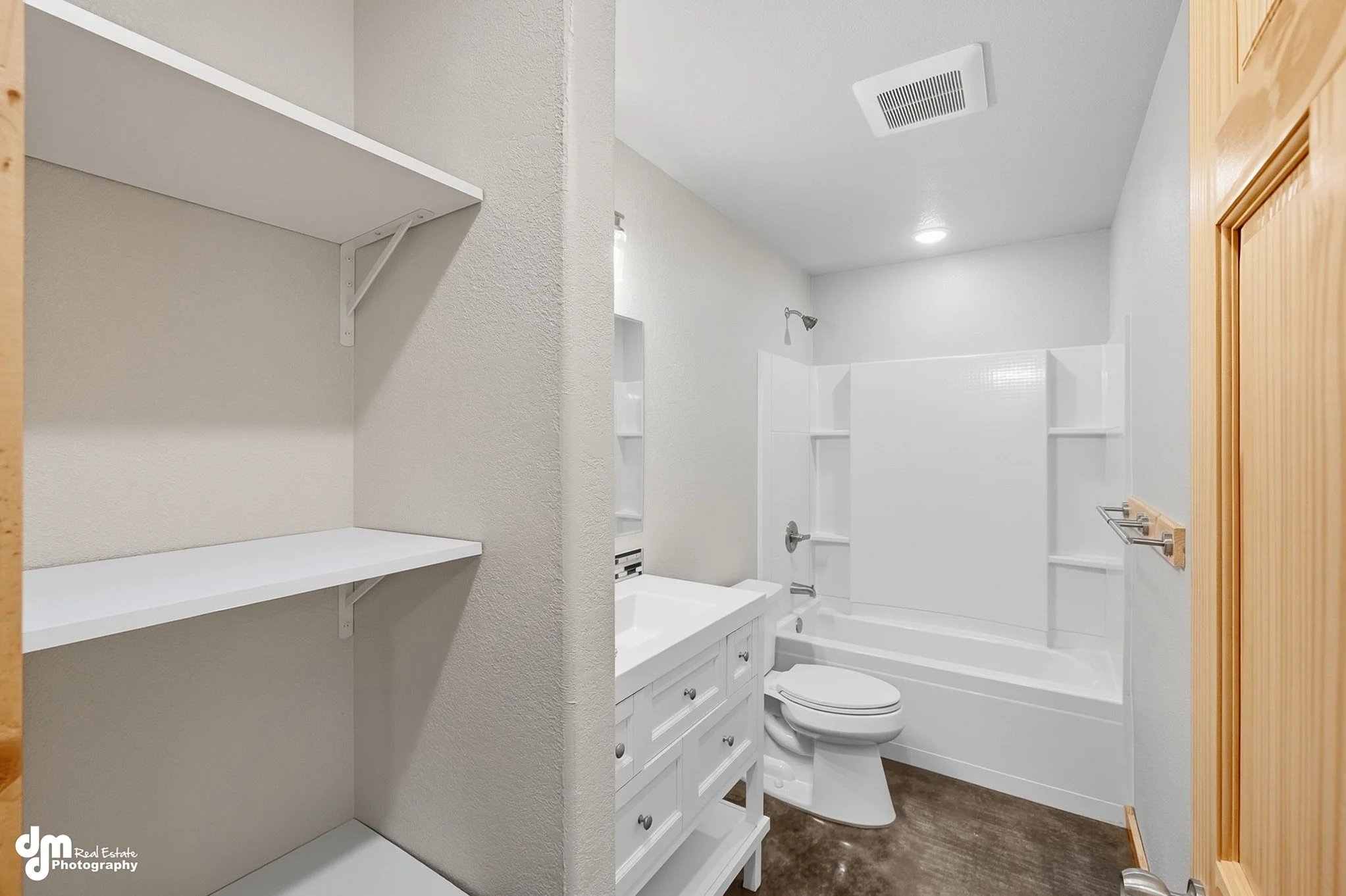 Bathroom with white bathtub and shower, toilet, white vanity with sink, and shelving, brown flooring, wooden door, and light-colored walls.