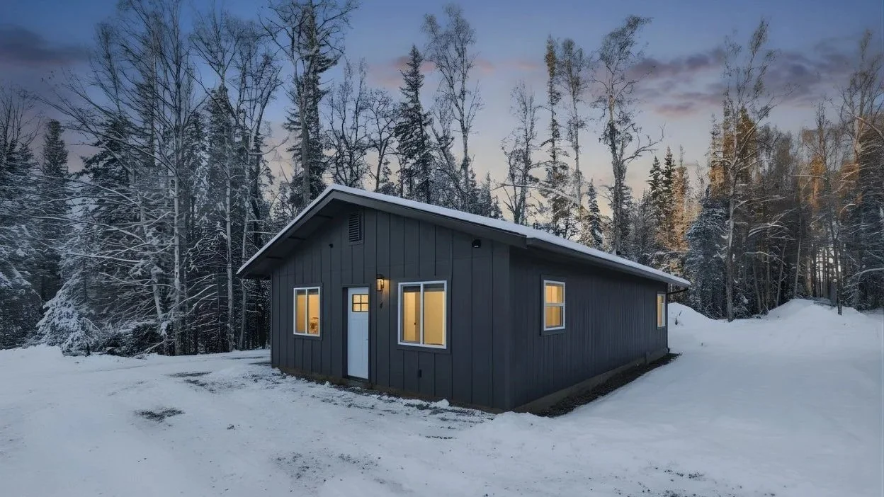 A small dark blue house with yellow-lit windows, set in a snowy landscape with trees in the background at dusk.