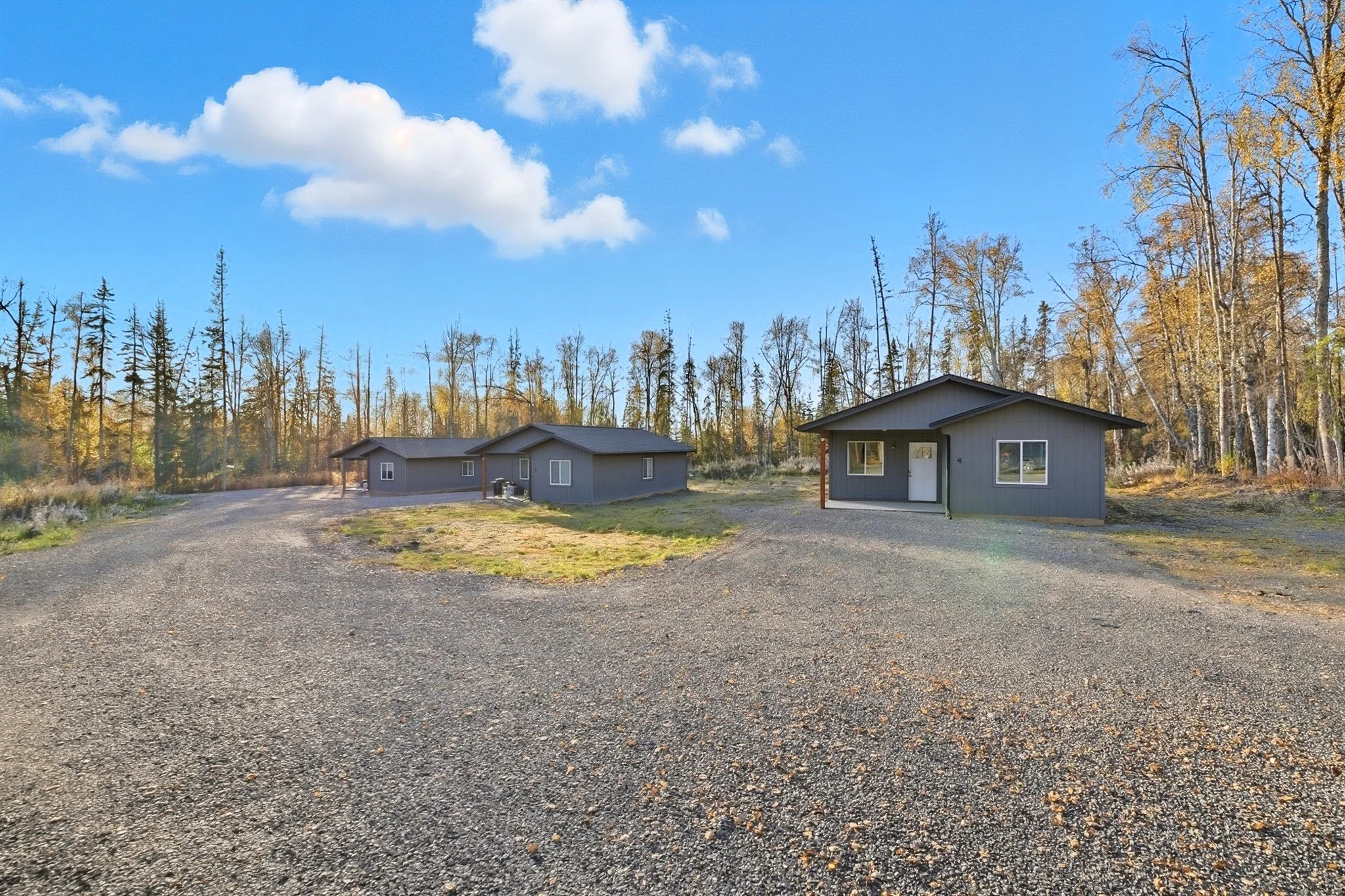 Three gray houses with white doors and windows situated on a gravel lot, surrounded by trees with fall foliage under a blue sky with white clouds.
