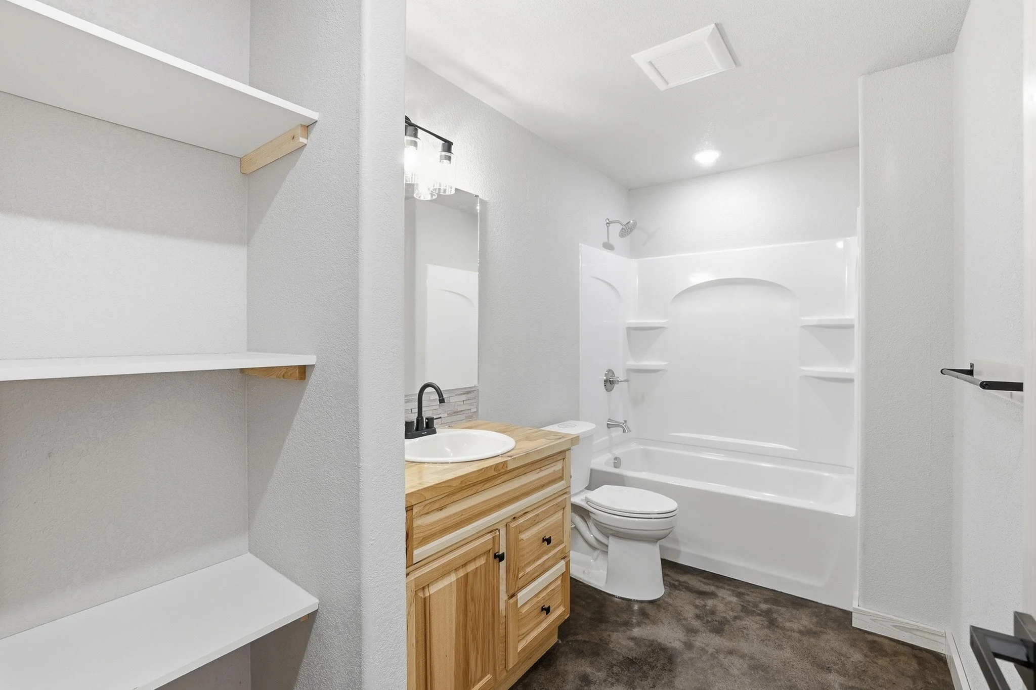 Bathroom with white walls, a wooden cabinet with a sink, a mirror, a toilet, a bathtub with shower, and white shelves on the left wall.