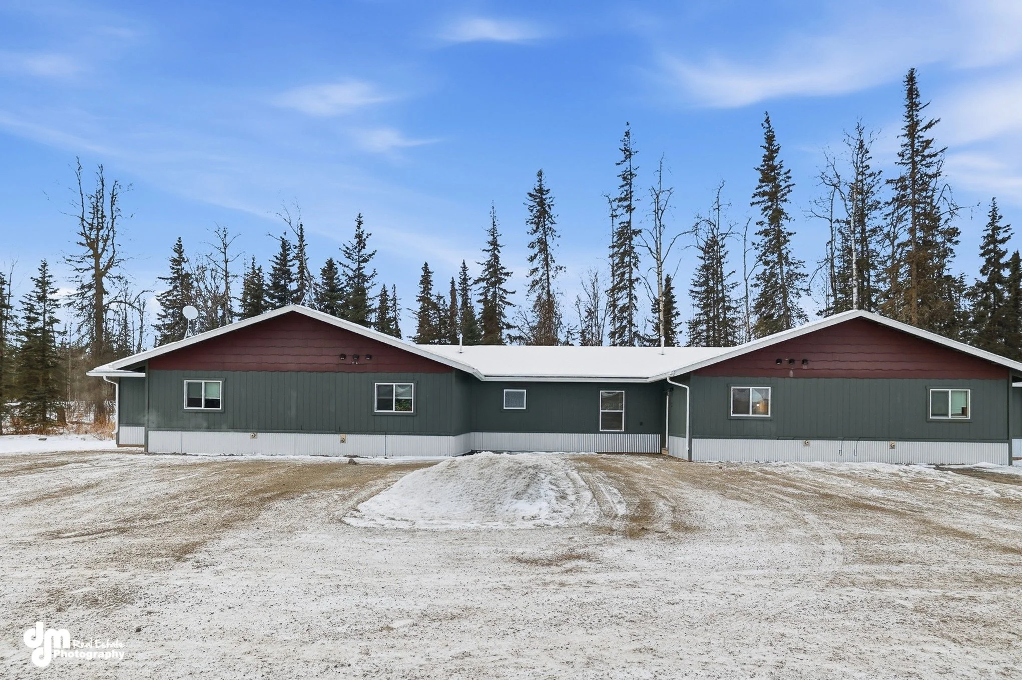 A single-story house with green siding, red gable ends, and a white roof, situated in a snowy landscape with tall, leafless trees in the background under a partly cloudy sky.
