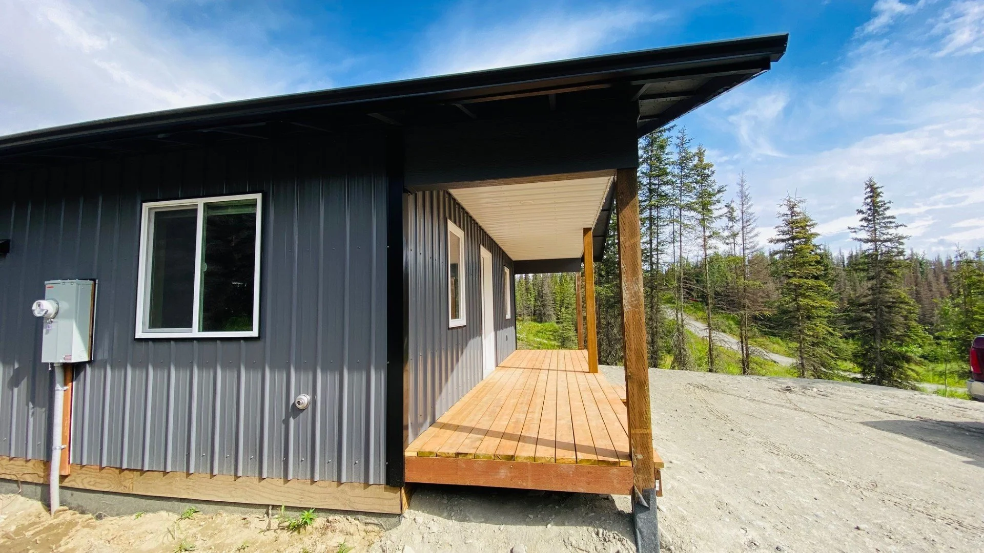 Front view of a modern house with black metal siding and a small wooden porch, surrounded by trees under a partly cloudy sky.