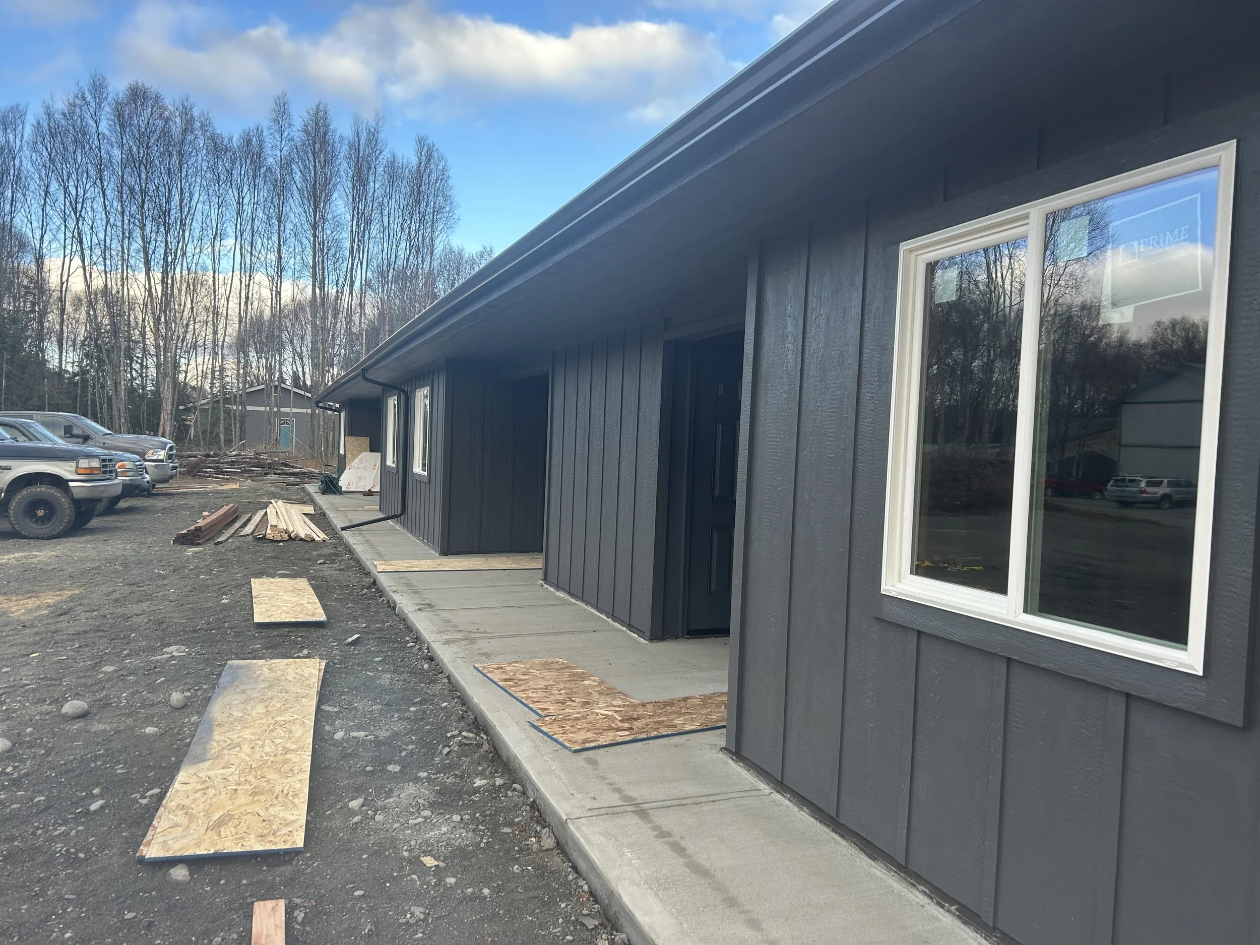 Construction site with a partially completed dark gray house, a concrete pathway, and wooden planks on the ground. Several parked vehicles are visible in the background. The sky is blue with clouds, and there are leafless trees around.