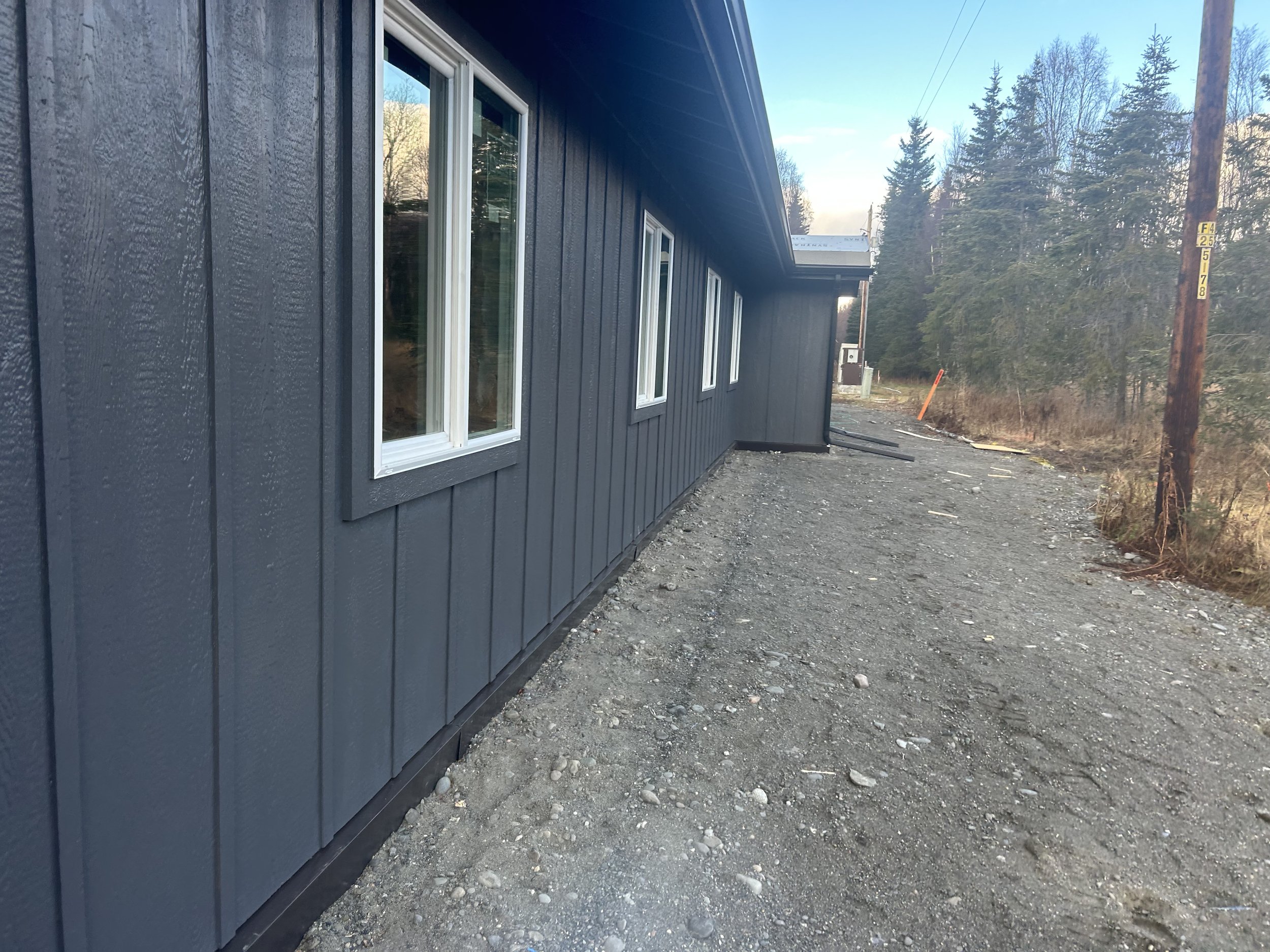 Side view of a newly constructed building with dark blue vertical siding, white window frames, and a gravel pathway next to it, in a rural area with trees and power lines in the background.