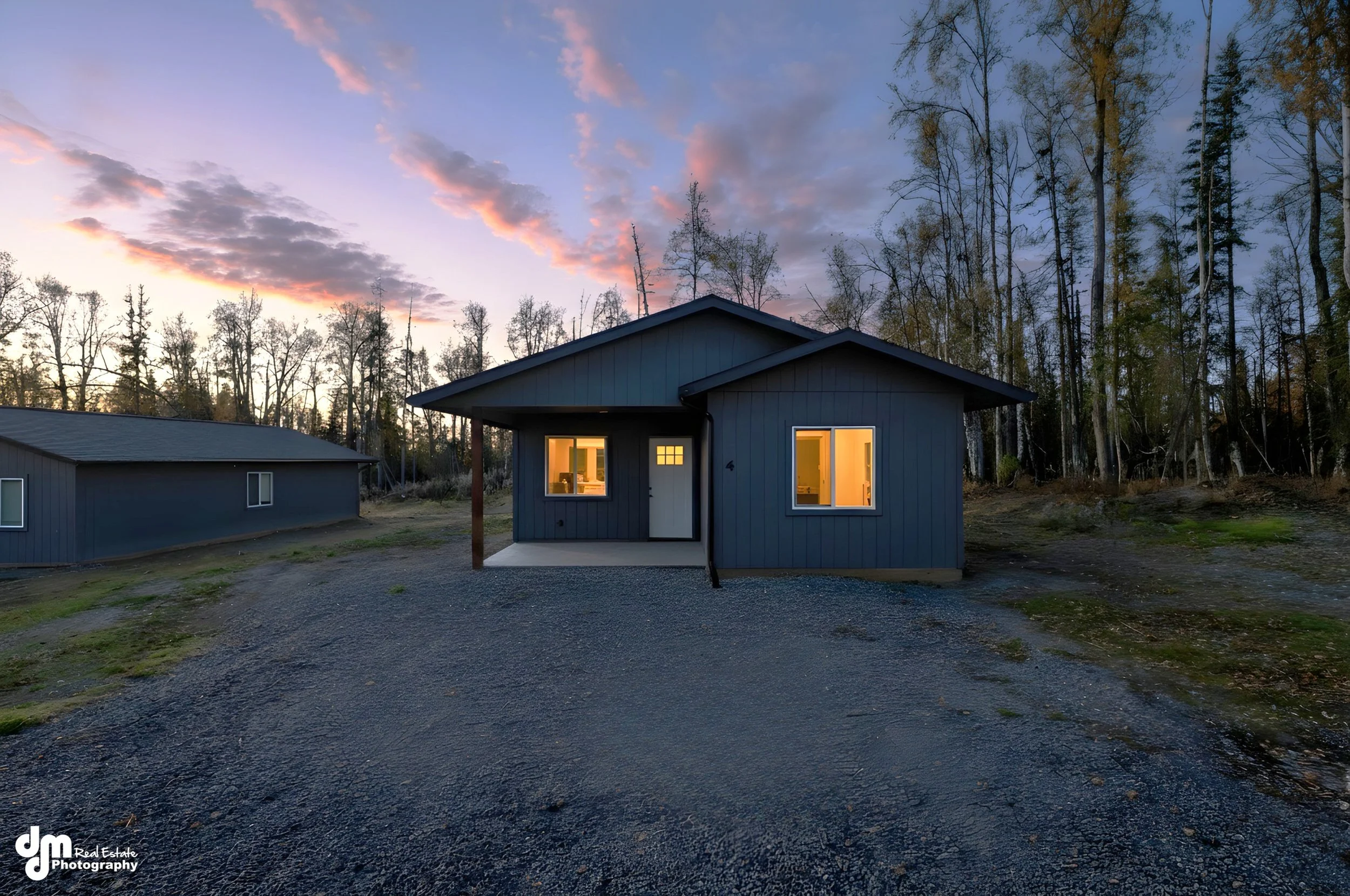 A small, blue house with illuminated windows during sunset, surrounded by trees and a gravel driveway.