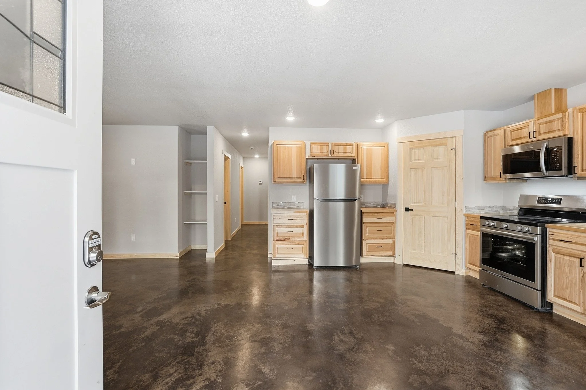 Empty kitchen with wooden cabinets, stainless steel refrigerator, oven, microwave, and dark polished concrete floor.