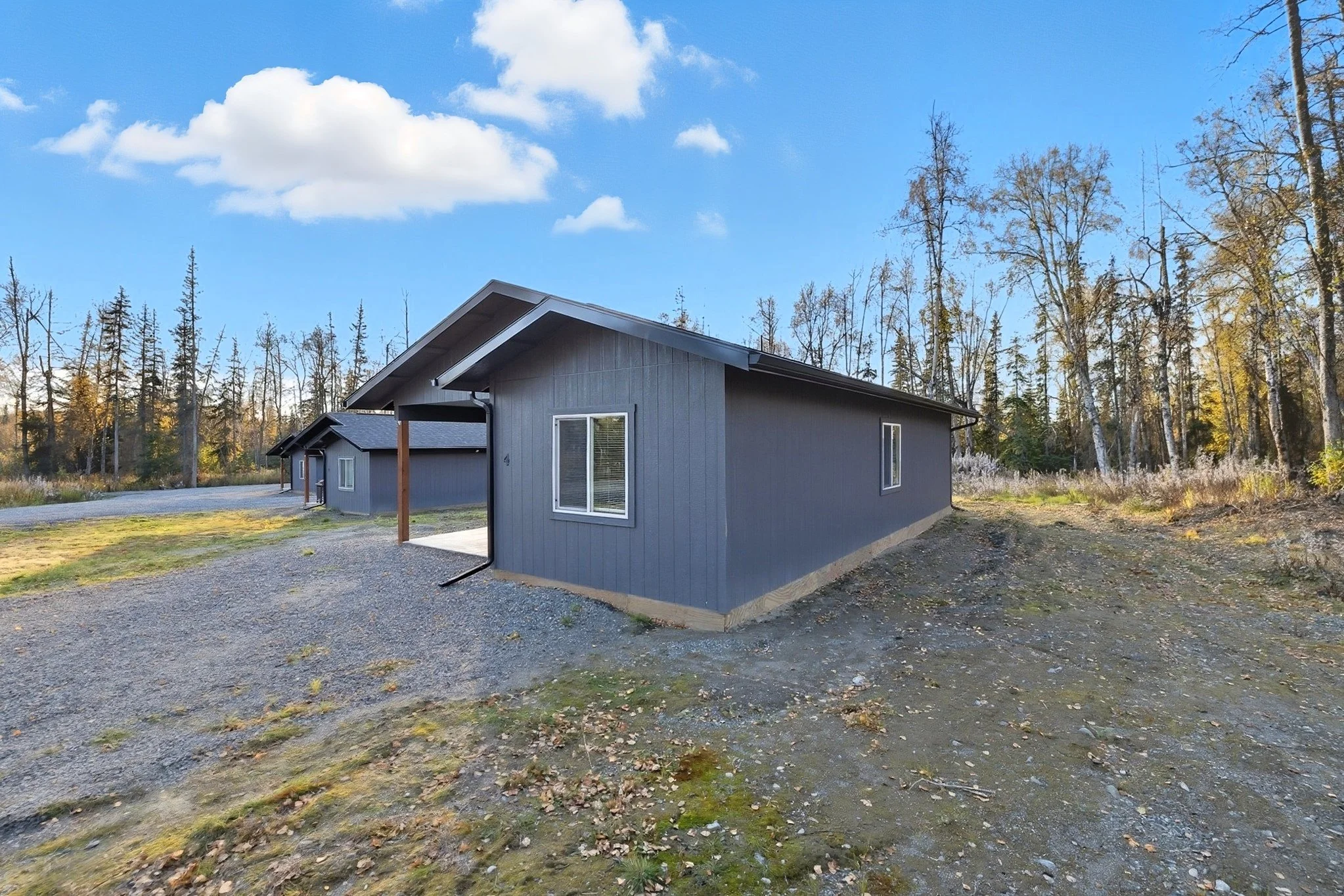 A single-story gray house with white trim in a wooded area, under a partly cloudy sky.