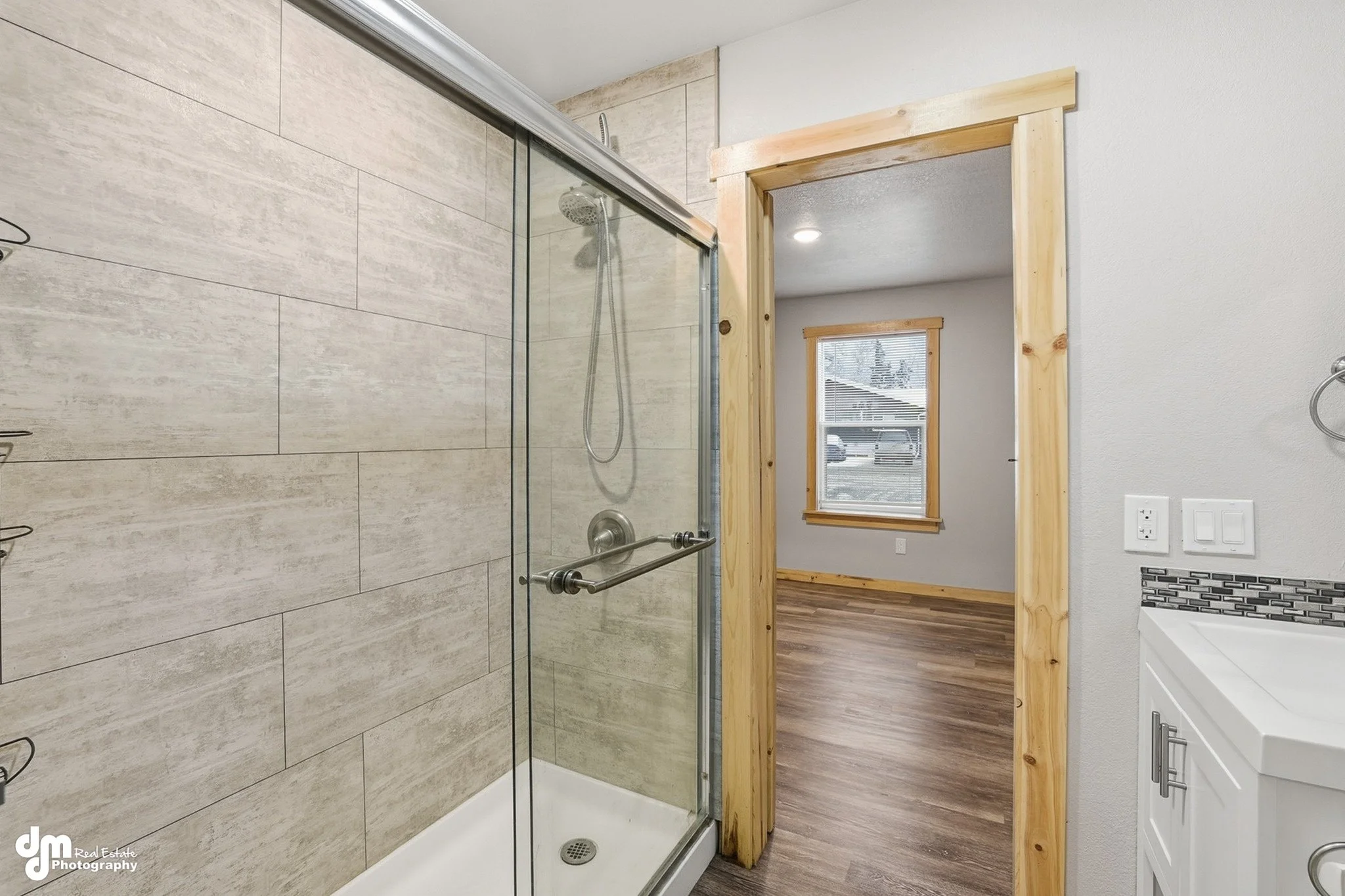 Inside a bathroom showing a glass-enclosed shower with beige tiles on the wall, a wooden doorframe, a window in the adjacent room, and part of a white vanity with a decorative backsplash.