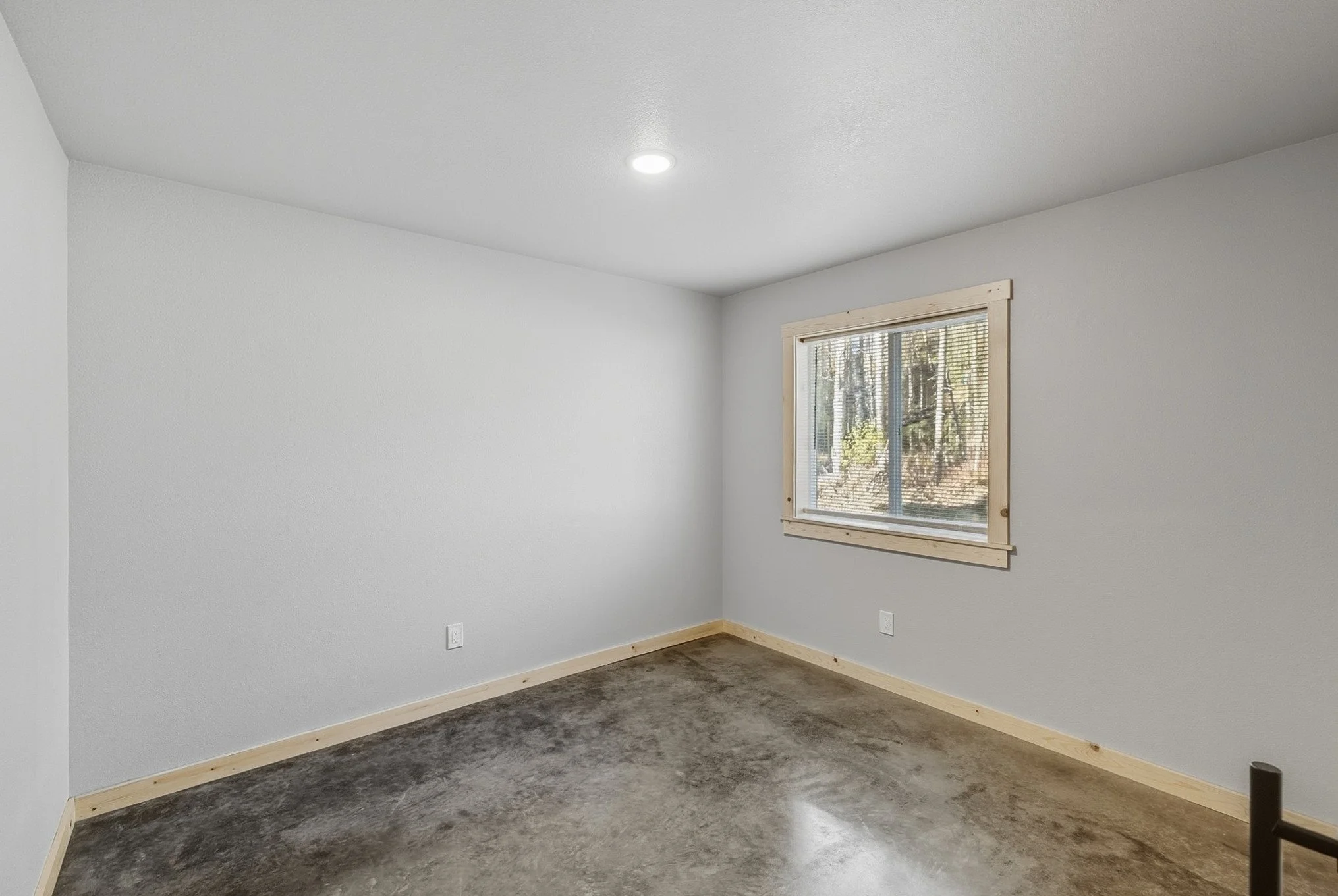 Empty room with white walls, a window with blinds showing trees outside, and a concrete floor.