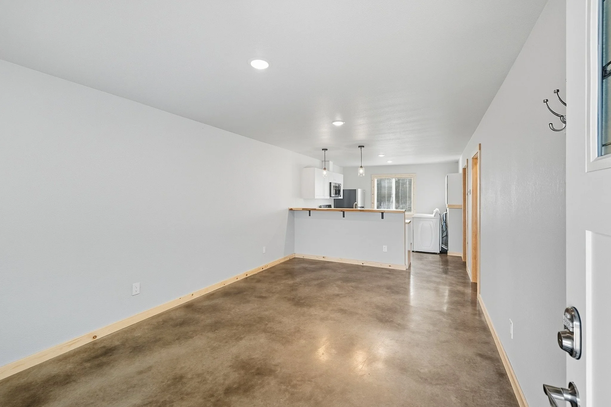 Empty living room with polished concrete floor, white walls, and a view into the kitchen area with pendant lights, a window, and laundry appliances.