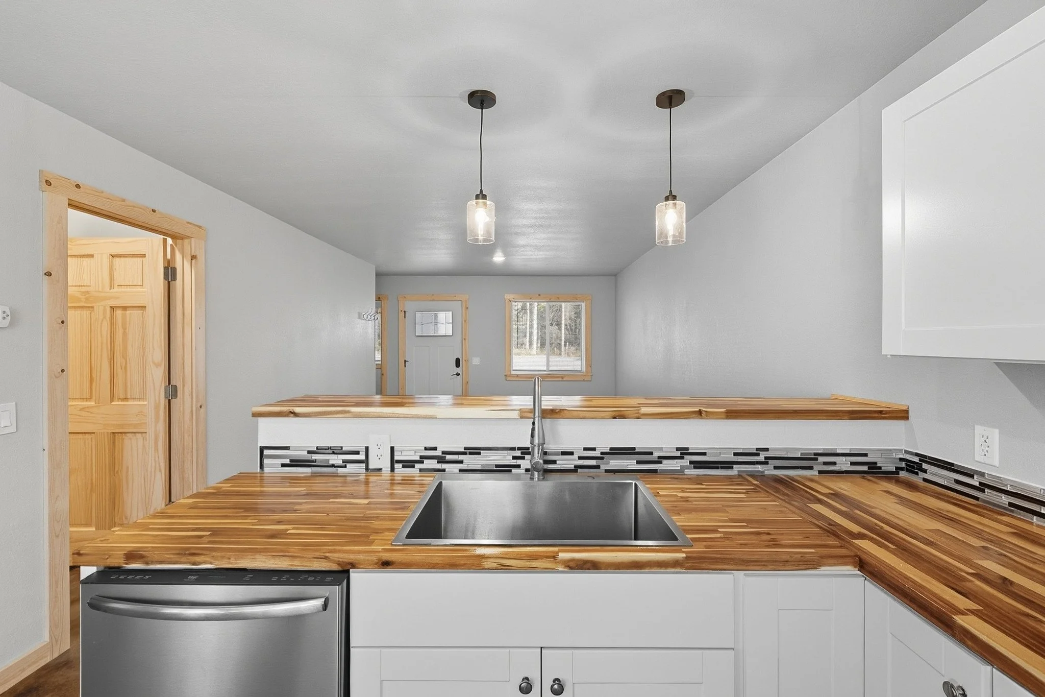 View of a modern kitchen with a wooden countertop, stainless steel sink, gray backsplash, white cabinets, and pendant lights hanging from the ceiling.