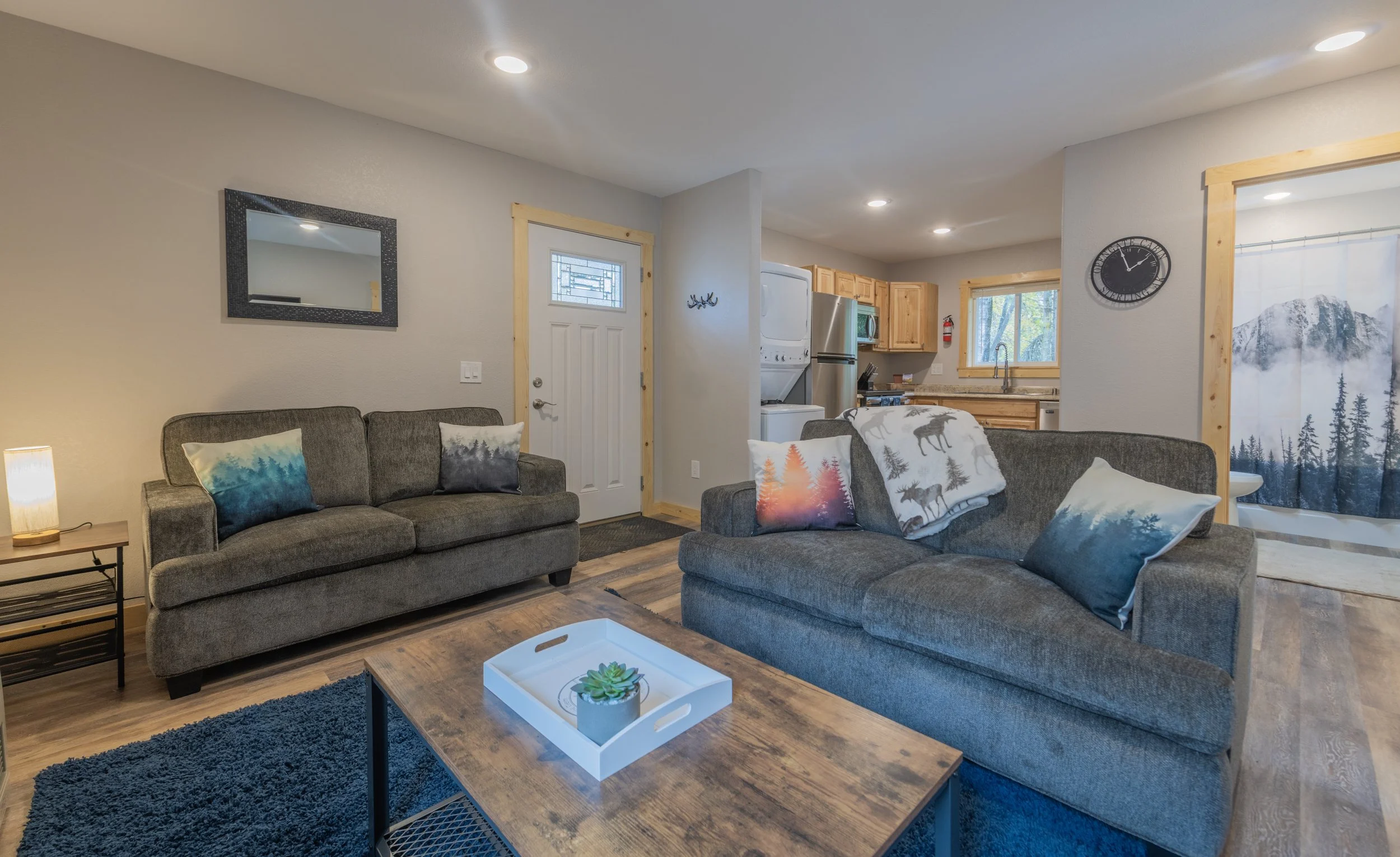 Living room with gray sofas, rustic wooden coffee table, and mountain-themed pillows, open to a kitchen and a doorway to a bathroom with mountain landscape curtain.