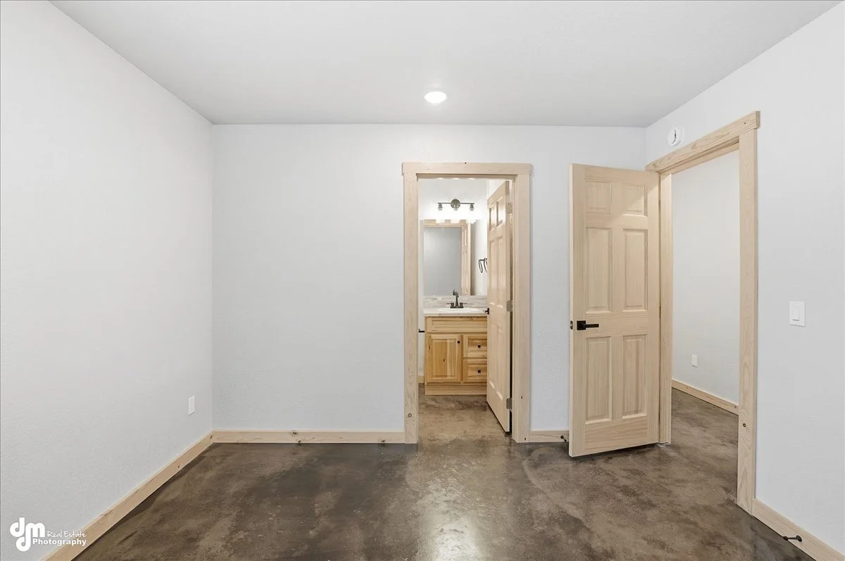 Empty room with white walls, natural wood trim, and a concrete floor. There are two doorways, one leading to a small bathroom with a sink and mirror, and another opening to a hallway. The room is lit by a ceiling light.