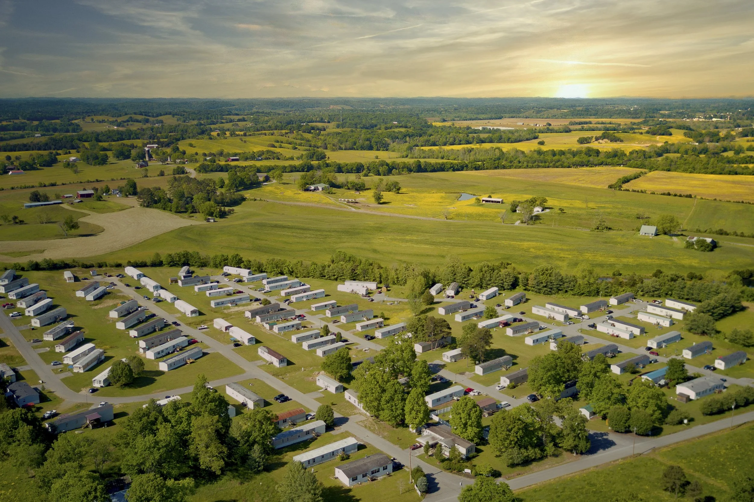 Aerial view of a rural landscape at sunset with a community of mobile homes, green fields, trees, and distant farmland.