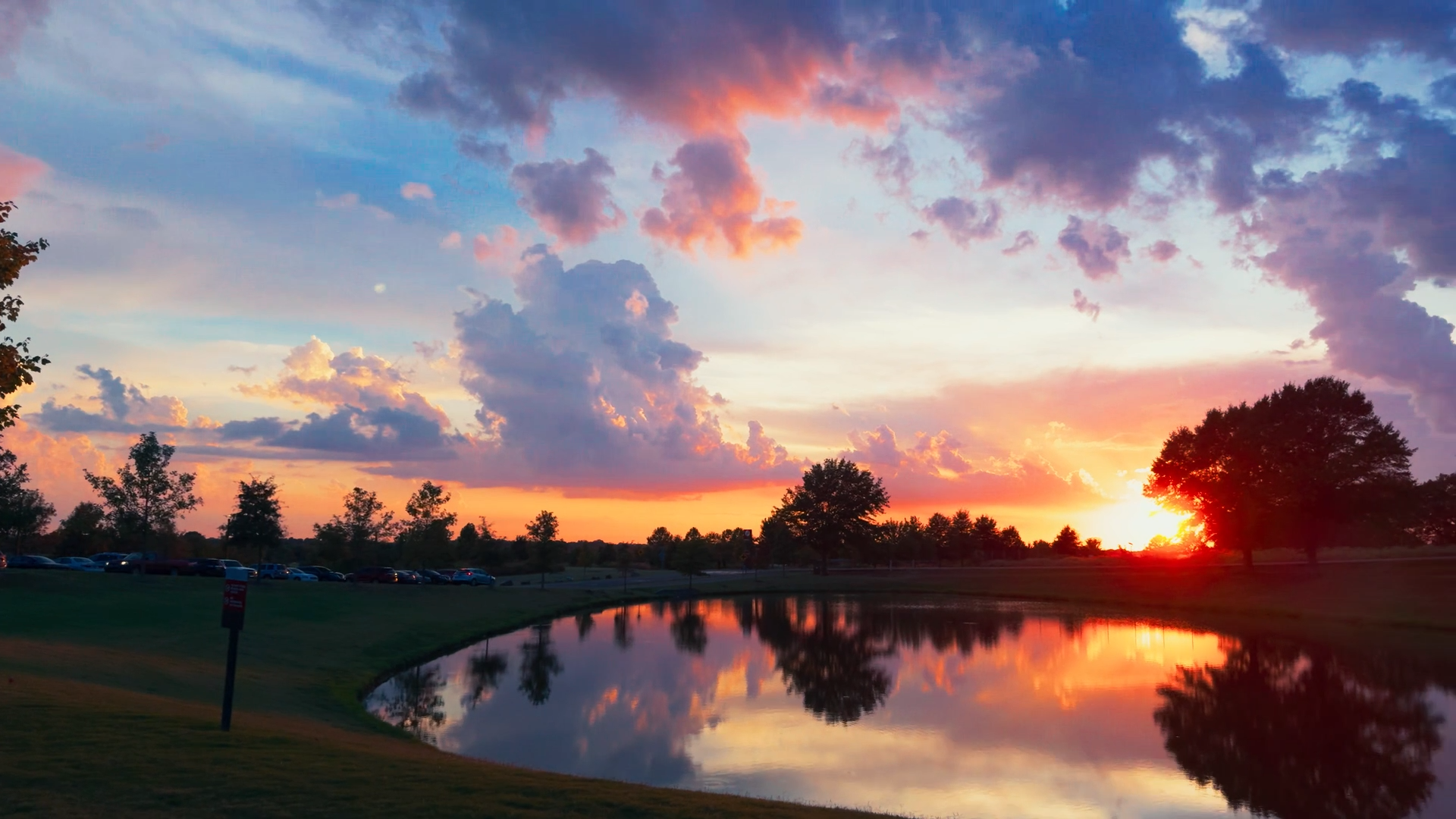 Beautiful sunset over a pond with reflection, surrounded by trees and a grassy area, partly cloudy sky with shades of pink, purple, and orange.