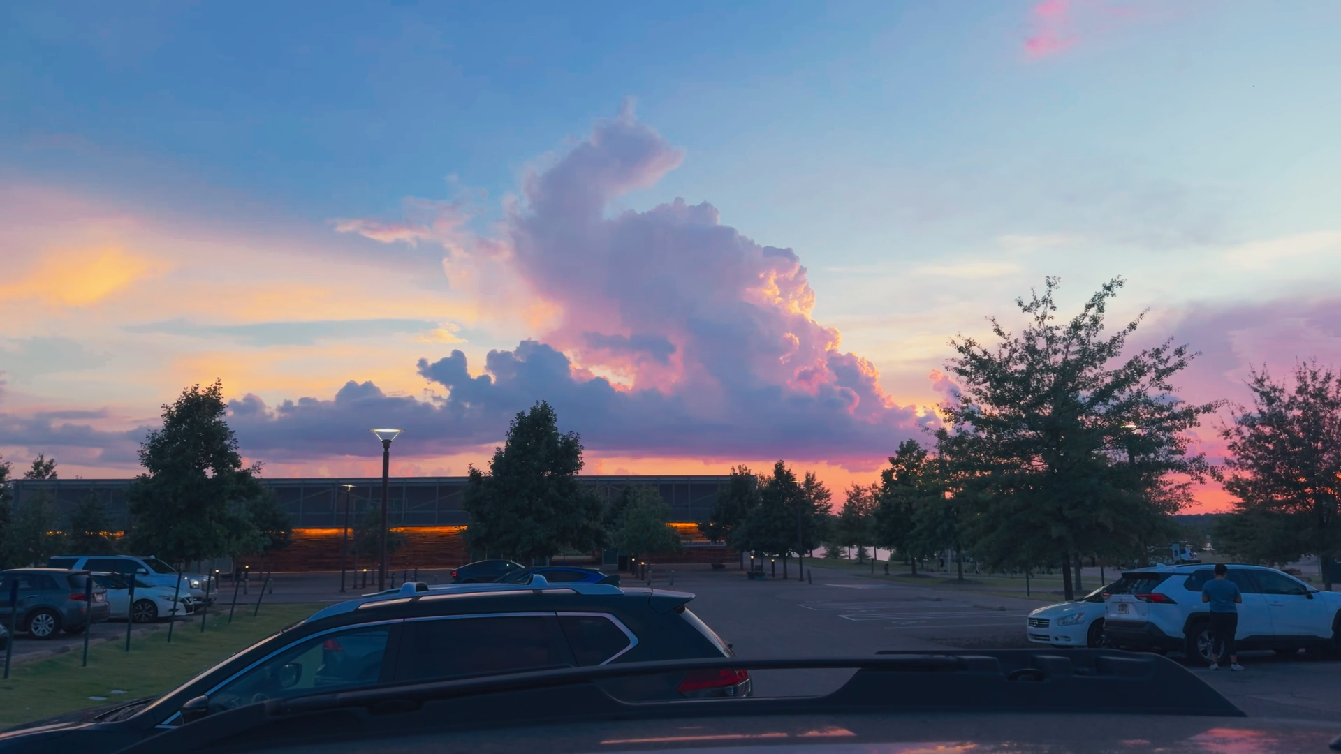 Beautiful sunset sky with large fluffy clouds over a parking lot with trees and people in the background.