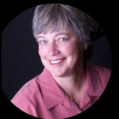 A woman with short gray hair wearing a pink collared shirt, smiling at the camera, against a dark background.