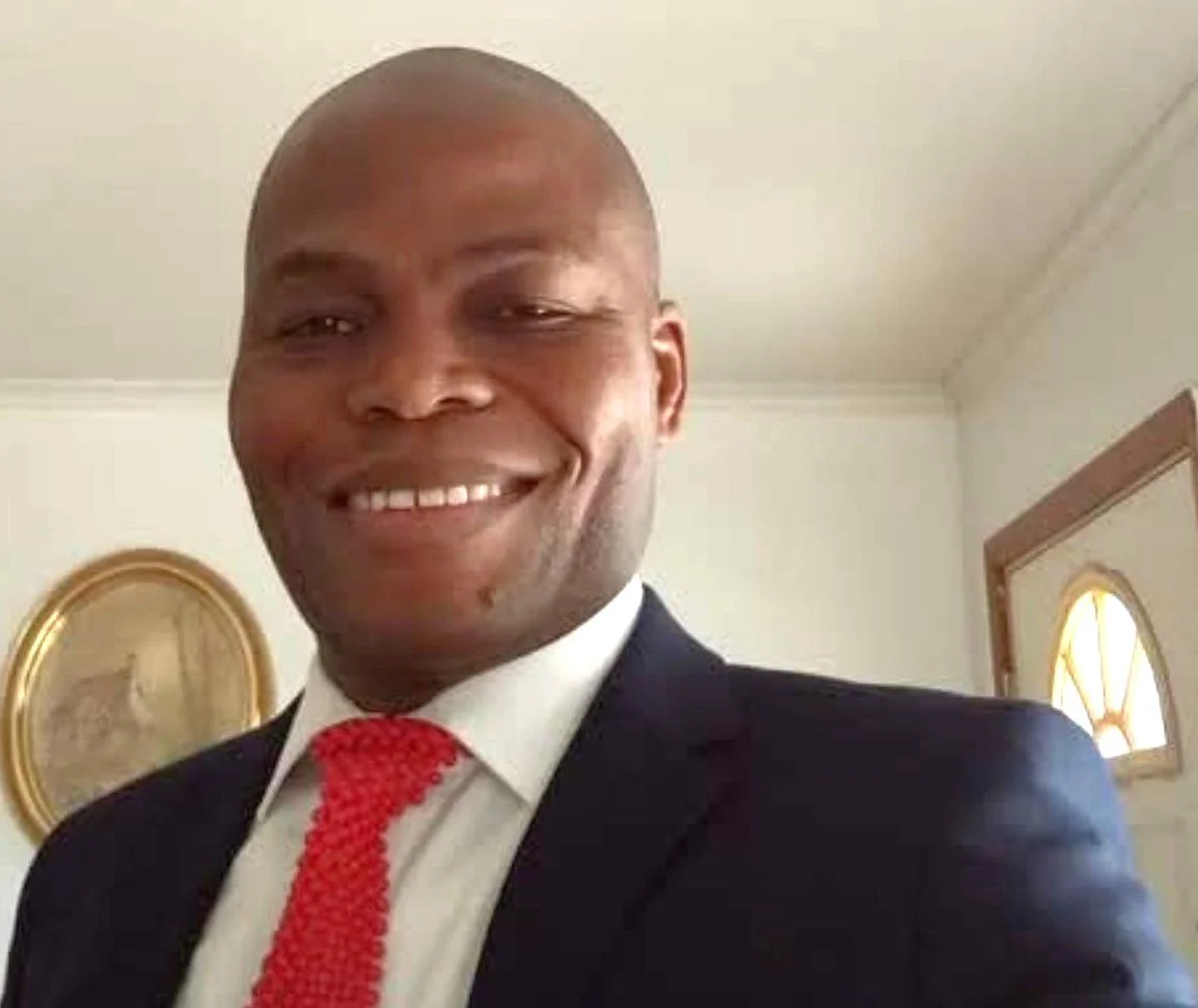A smiling man wearing a dark suit, white shirt, and red tie, standing indoors with a gold-framed picture and an arched window visible in the background.