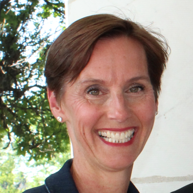 A smiling woman with short brown hair standing outdoors with trees and blue sky in the background.