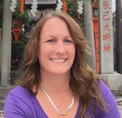 A smiling woman with wavy brown hair wearing a purple top and pearl necklace, standing in front of a traditional Japanese shrine with red torii gates and stone statues.